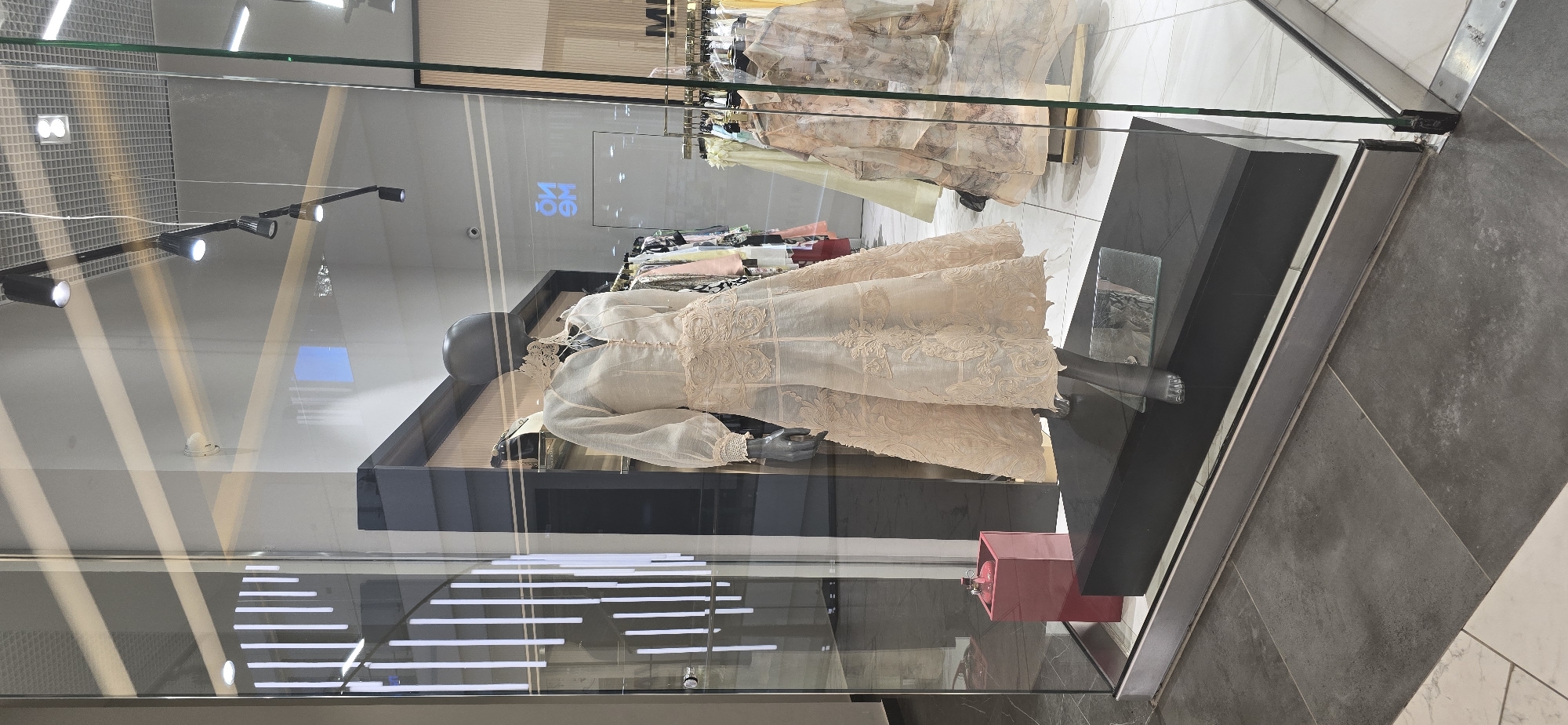 The image shows a mannequin in a glass display case at a retail store in Grozny, Russia. The mannequin wears a cream-colored dress with lace detailing. The display case sits on a dark gray tile floor. In the background, racks of clothing are visible. Lighting fixtures are mounted on the ceiling. The storefront features a reflective glass facade.