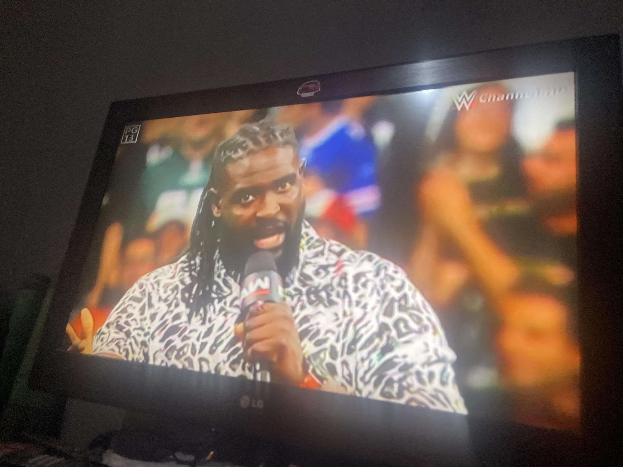 The image shows a television screen displaying a wrestling event. A man with dark skin and braided hair is speaking into a microphone. He is wearing a white shirt with a black, abstract pattern. Behind him, a crowd of people is visible, with some in bright colors. In the top left corner of the screen, there is a "PG 13" rating. In the top right corner, a "WWE Channel HD" logo is visible. The television screen itself is framed by a black bezel, and the bottom bezel has an "LG" logo. The overall lighting suggests the room is dimly lit. There is no information in the image to suggest the location is the city of Jos, Nigeria.