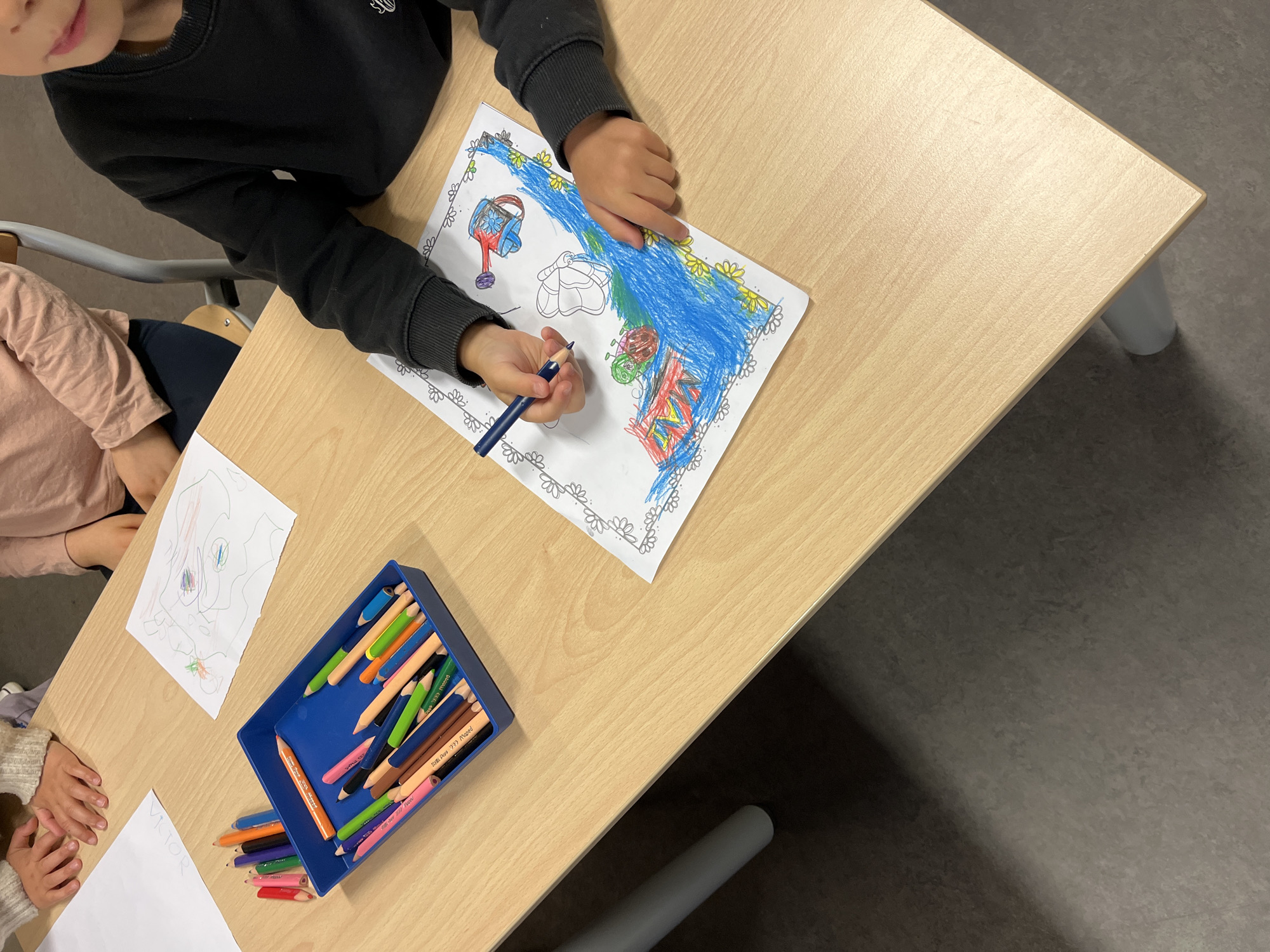 The image depicts a child coloring at a light-colored wooden table. The child, wearing a black long-sleeved shirt, is focused on coloring a picture of a garden scene with a watering can, flowers, and other objects. A box of colored pencils sits on the table next to the drawing. Another child, wearing a pink long-sleeved shirt, is partially visible on the left side of the image with a blank sheet of paper and another drawing on the table next to them. The setting is an indoor space, likely a classroom, located in La Fouillouse, France.