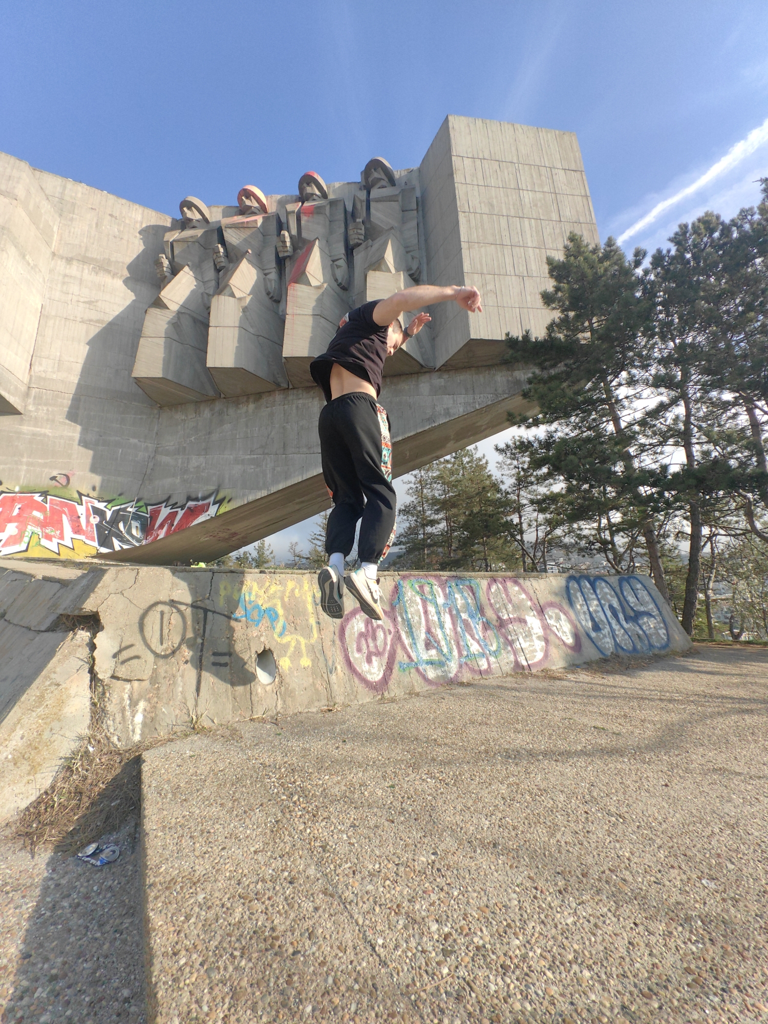 A male individual is captured mid-jump in front of the Monument to the Bulgarian-Soviet Friendship in Varna, Bulgaria. The subject wears a black short-sleeved t-shirt, black sweatpants with a visible colorful pattern on the right leg, white socks, and white sneakers with black accents. His arms are bent and raised, and both feet are off the ground, indicating an athletic movement.
The background is dominated by the monumental brutalist concrete structure, featuring multiple stylized, abstract figures projecting from its upper facade. Several of these figures show evidence of red spray paint on their upper sections. The lower part of the monument's base and a foreground concrete wall are extensively covered in multi-colored graffiti tags and markings, including red, green, yellow, blue, and white. The foreground consists of a gravelly concrete surface. Pine trees are visible to the right and behind the monument, under a clear blue sky with a faint contrail. Strong sunlight casts distinct shadows. Two metallic can pull-tabs are discernible on the ground in the lower-left corner.