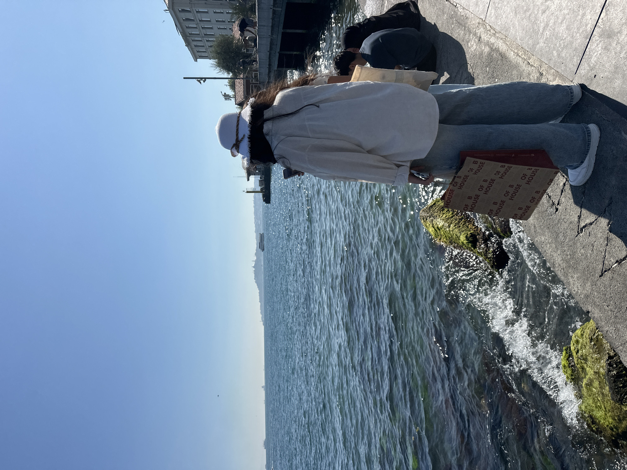 In Beşiktaş, Turkey, on a sunny day, a young woman stands on a concrete embankment overlooking the Bosphorus. She wears a white hat, a light-colored oversized shirt, and loose-fitting denim jeans, holding a reusable shopping bag with the "HOUSE OF B" logo. To her right, two individuals are seated on the embankment; one wearing a dark shirt and the other a black headscarf. The water is calm with gentle waves lapping against moss-covered rocks at the edge. In the distance, across the water, the silhouette of a cityscape with buildings and a minaret is visible under a clear blue sky, with several boats dotting the horizon. A pier with structures and trees extends into the water on the right side of the frame.