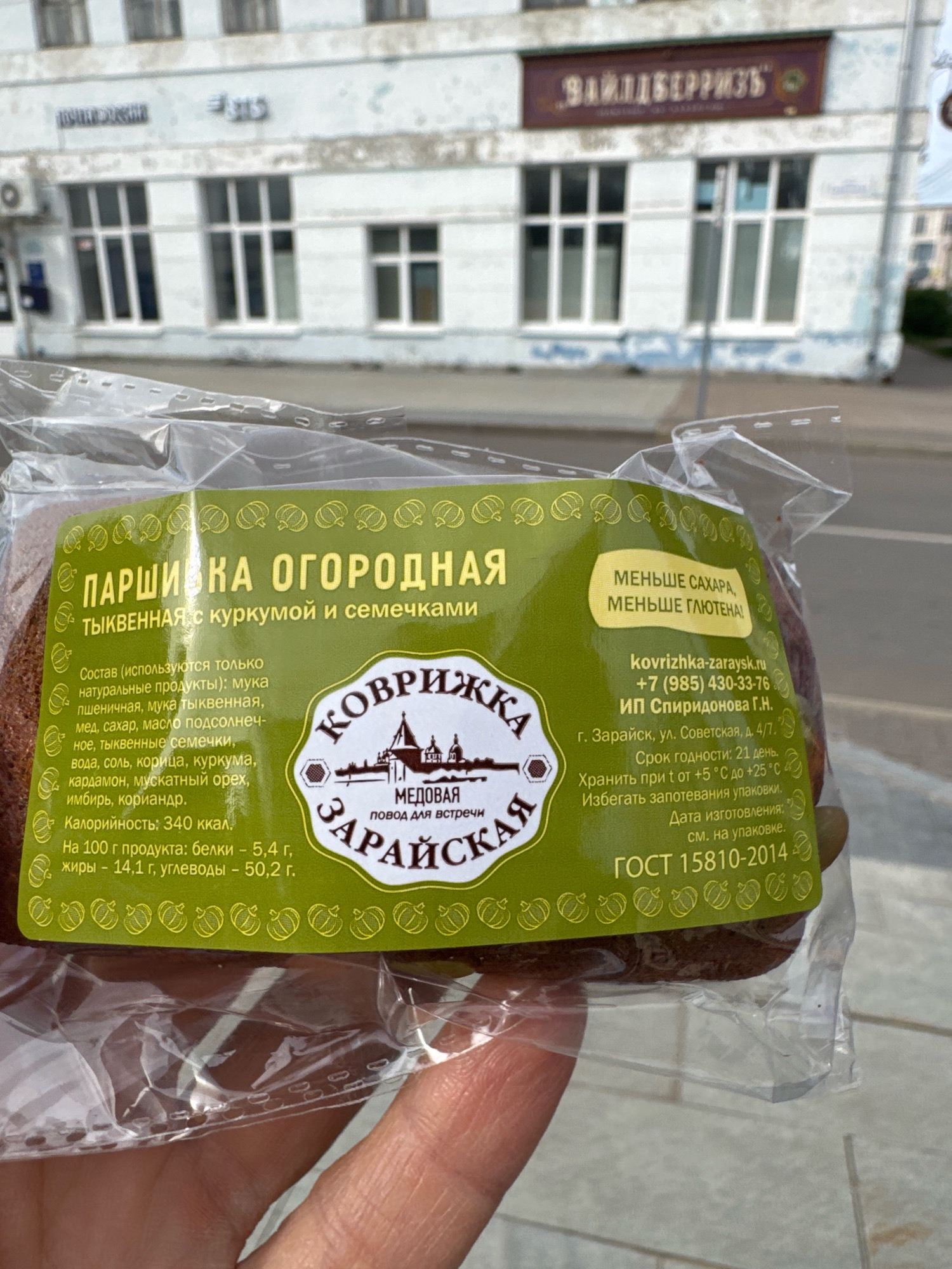 An outdoor, eye-level, slightly upward-angled shot shows a person's hand holding a package of baked goods, which appears to be a loaf of bread or a large cookie. The product is round and dark brown, wrapped in clear plastic. The label is green and features text and images in yellow and white. The text indicates it is "PARSHIVKA ORODNNAYA" (likely "Garden Parsley" or "Garden Rye") and "TYKVENNAYA s kurkumoy i semechkami" (Pumpkin with turmeric and seeds). It also lists ingredients, nutritional information, and manufacturer details.
The manufacturer's information states it is from Zaraysk, Ul. Sovetskaya, d. 4/7, and provides a website, phone number, and individual entrepreneur name. The background is blurred but shows a light-colored building with multiple windows, suggesting an urban or town setting. Above the building, a sign reads "BAILLIBERIZ" in dark red letters on a brown background, possibly indicating a business name. Another partial sign on the left reads "ПОЧТАРОССИИ" (Russian Post). The overall environment appears to be a sunny day.