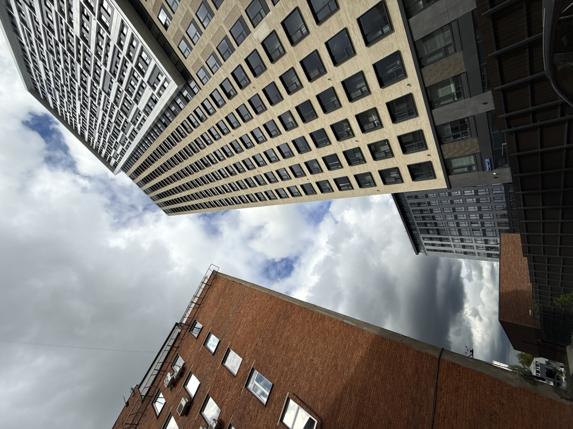 A low-angle shot looks upward at several buildings in Khimki, Russia. The perspective emphasizes the height and converging lines of the structures against a cloudy sky.
Four buildings are visible in the frame. The building at the bottom is a reddish-brown brick structure with multiple windows. To the right is a taller modern building with a beige-colored facade with repetitive window patterns and vertical architectural elements. The sky is partially visible between the buildings, showcasing a mix of light and dark clouds. A building in shades of gray is seen on the top left. Another more modern-looking gray building is on the top right.