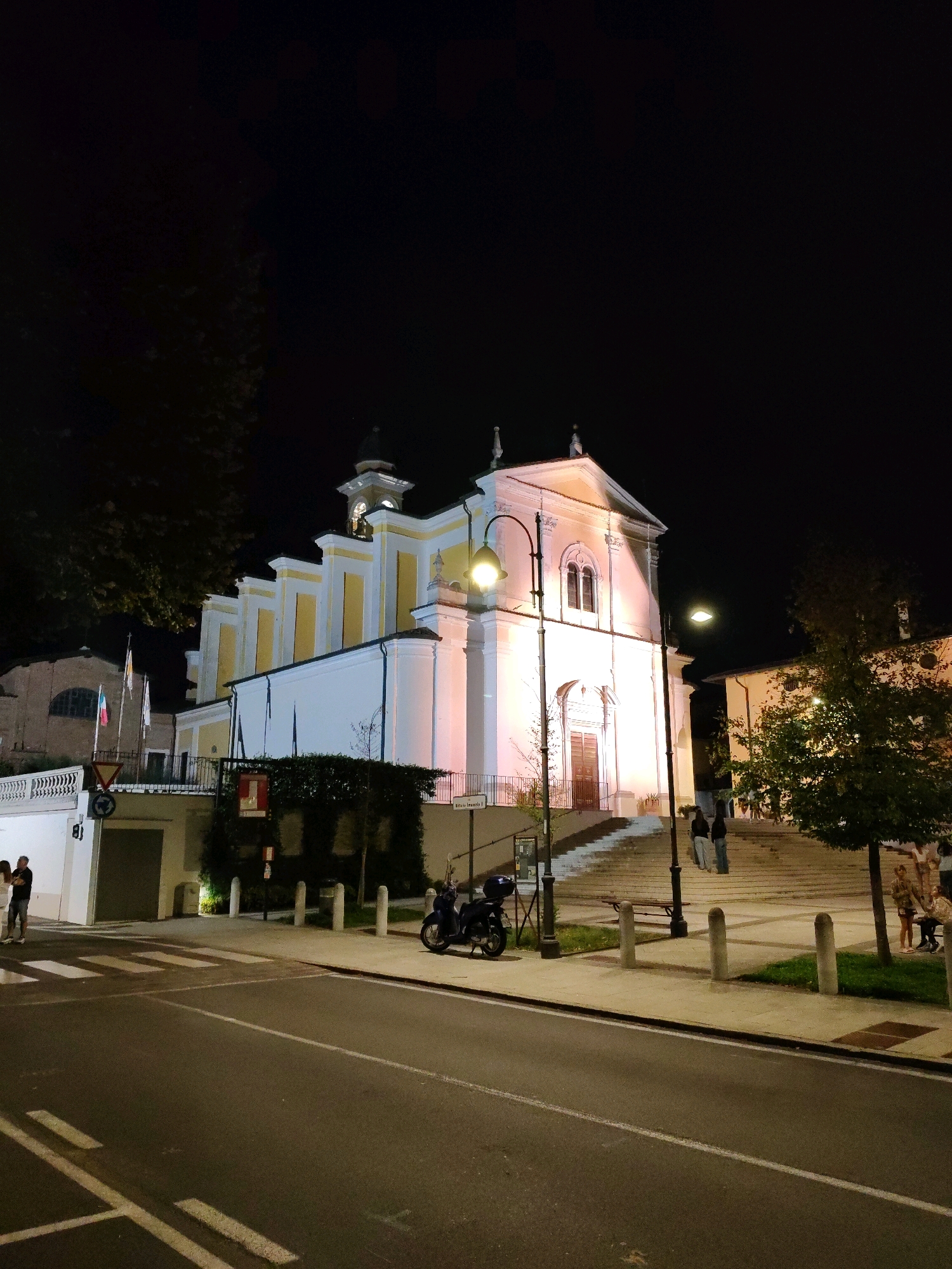 A nighttime exterior shot features a brightly lit church with white and yellow facades. The church has a prominent bell tower with a clock and a peaked roof. A set of wide stairs leads up to the main entrance. A street with a crosswalk is visible in the foreground, with a parked scooter on the sidewalk. Several people are gathered near the church and on the stairs. Trees and streetlights are also present, with the sky appearing completely dark. The scene is illuminated by artificial lighting.