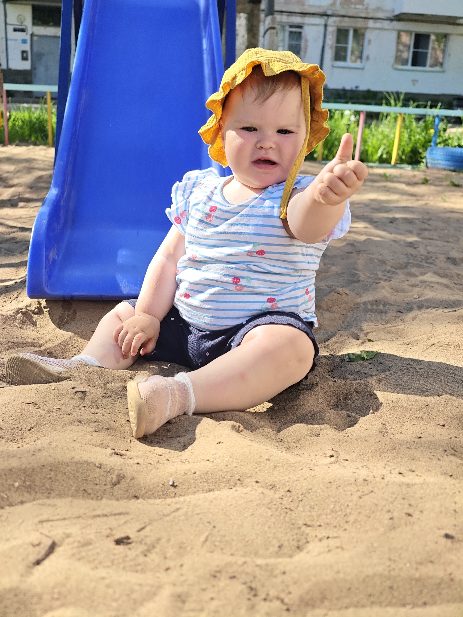 A baby sits in a sandbox in Chaikovsky, Russia. The baby wears a yellow sun hat, a short-sleeved shirt with blue and white stripes and red cherries, and navy-blue shorts. The baby gives a thumbs-up with the right hand. A blue plastic slide is next to the child on the left. A building with a light facade is in the background, along with grass and trees. The sandbox is in a playground area with colorful fencing and a tire swing.