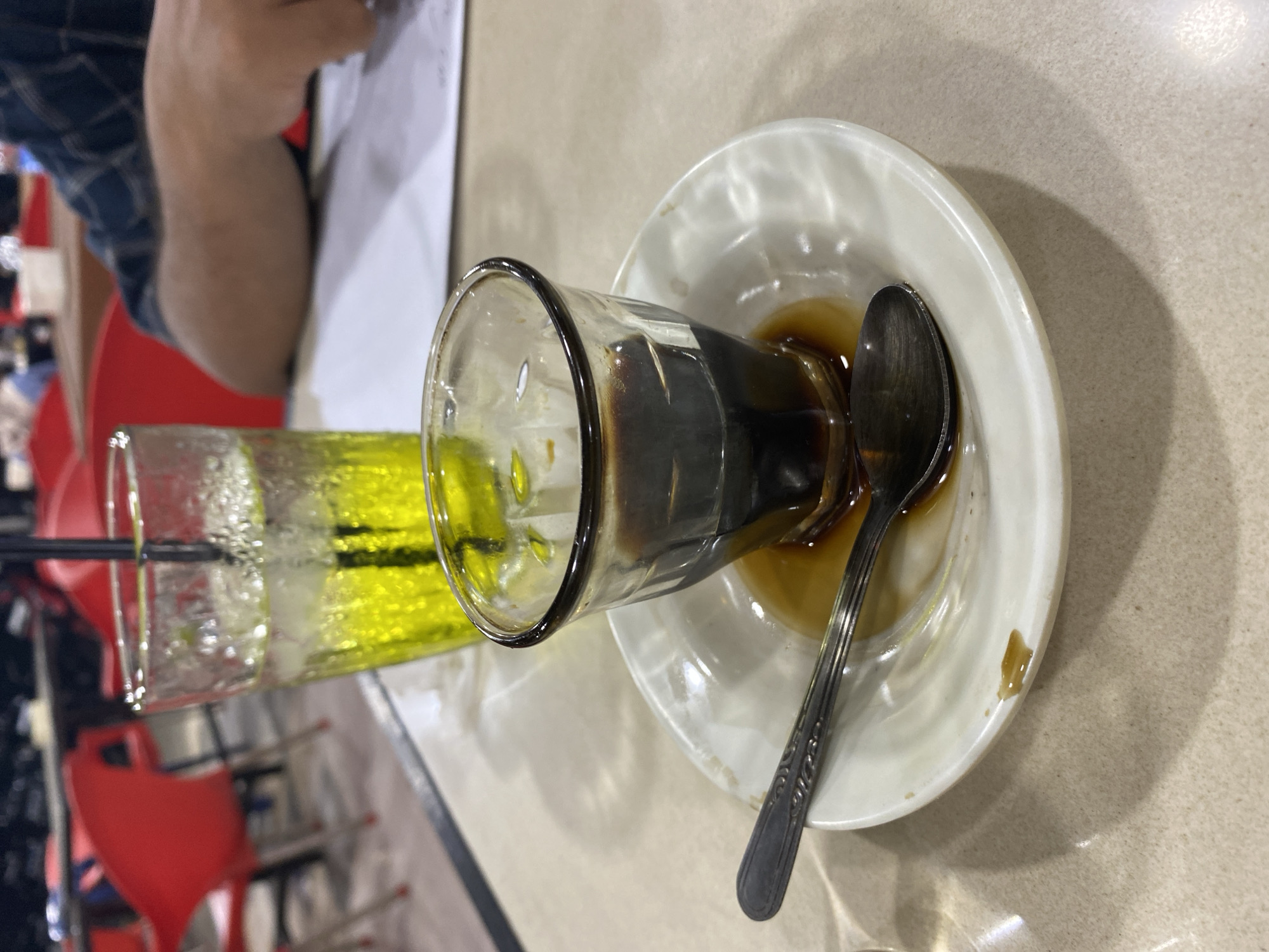 The image shows a close-up view of a small glass of dark liquid, likely coffee or espresso, on a white saucer. A spoon rests on the saucer, partially submerged in a small amount of spilled liquid. In the background, a tall glass filled with a bright yellow, bubbly beverage, possibly soda or iced tea, with a black straw is visible. The setting appears to be a restaurant or cafe, with red chairs and tables in the background, suggesting an indoor environment. The presence of papers on a table in the background hints at a casual dining or meeting space. There are no clear identifiers of the location within Lhokseumawe, Indonesia, but the general ambiance suggests a public establishment.