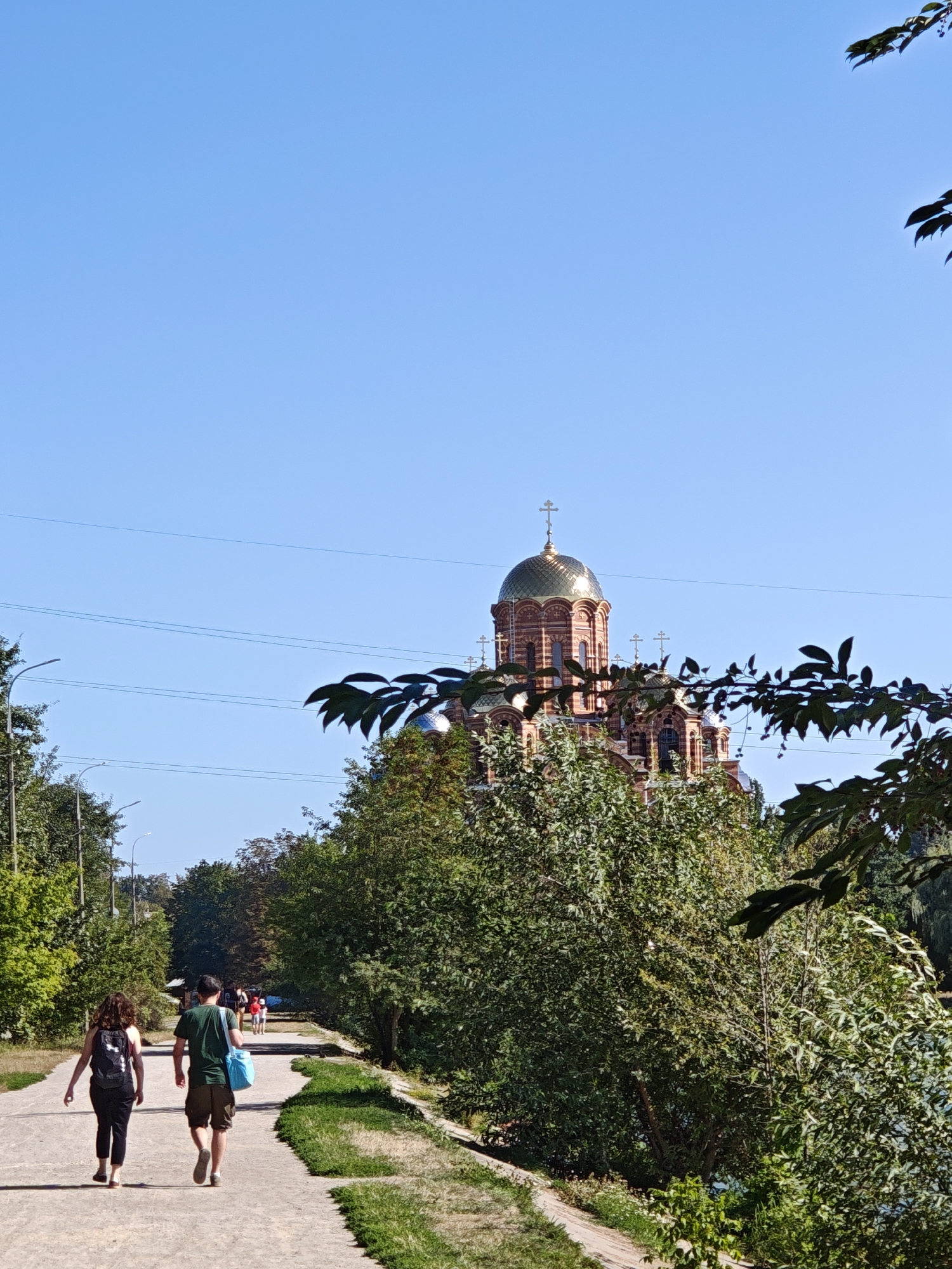 In Vinnytsia, Ukraine, on a sunny day, a paved pathway runs through a park-like setting lined with trees. A woman and a man walk away from the camera on the pathway, with the woman wearing a black tank top and black leggings and carrying a black backpack, while the man wears a green t-shirt and khaki shorts and carries a blue bag. In the background, partially obscured by trees, is a large brick church with a golden dome. The sky is clear and blue.