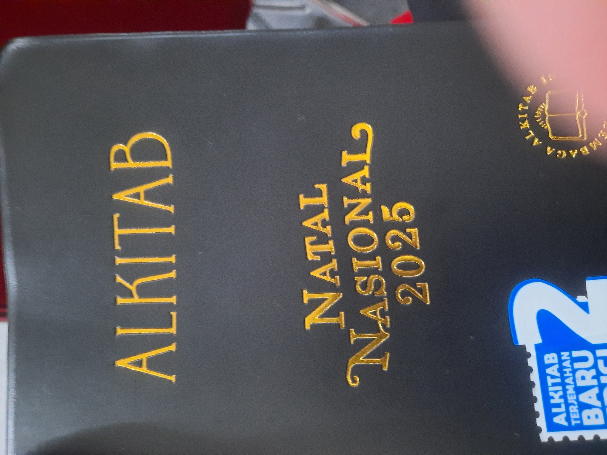 The image shows a closed black Bible with gold lettering. The text on the cover reads "ALKITAB" in large letters at the top, followed by "NATAL NASIONAL 2025" in slightly smaller, decorative script. At the bottom left, there is a blue and white sticker with text that includes "ALKITAB TERJEMAHAN BARU". To the bottom right, there is a circular gold emblem with text and an open book icon. The background is dark and appears to be an indoor setting, with hints of red and white elements in the upper right corner. The lighting is direct, creating some glare on the glossy surface of the Bible. The image does not provide any information about the location being North Jakarta, Indonesia.