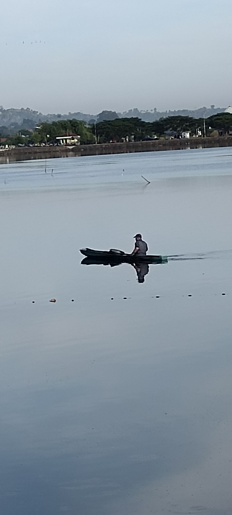 A lone individual, wearing a dark cap and shirt, sits in a dark-colored, narrow boat, likely a kayak or canoe, on a calm body of water. The boat is positioned horizontally in the middle of the frame, leaving a long, dark reflection on the water's surface. The individual appears to be facing towards the right side of the image, with their posture suggesting they are actively paddling or resting. The water is a muted blue-gray, reflecting the overcast sky. In the background, a low-lying shoreline is visible, characterized by a concrete wall topped with trees and some buildings, suggesting an urban or semi-urban environment. The distant hills are shrouded in a light mist, creating a soft, hazy effect. The overall scene evokes a sense of quiet solitude and tranquility. Based on the landscape and common settings in Lhokseumawe, Indonesia, this scene is likely captured at the Reservoir Lake or a similar body of water within the city.