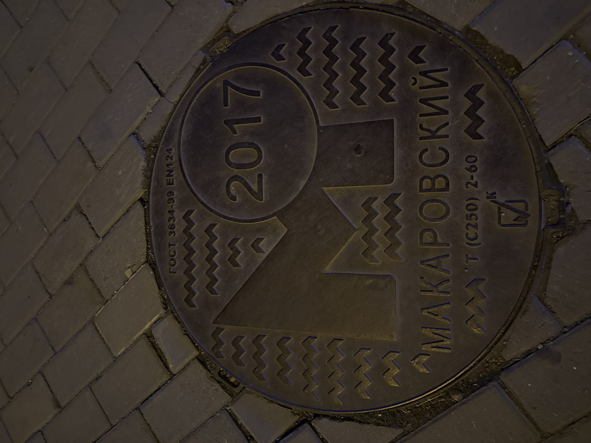 The image shows a close-up of a manhole cover embedded in a brick-paved surface. The manhole cover is circular and made of a dark metal, likely cast iron. The year "2017" is prominently displayed in a circle on the upper left quadrant of the cover. A stylized, geometric design resembling a mountain or a building facade occupies the central area. Decorative zigzag patterns frame the central design. Cyrillic text, "МАКАРОВСКИЙ," arcs across the upper right portion of the cover. Additional text below it includes "T (C250) 2-60". The lower right quadrant has a logo. "ГОСТ 3634-99 EN124" is printed on the upper left. The brick pavement surrounding the manhole cover is dark and wet. The scene appears to be in an urban setting, likely a street in Yekaterinburg, Russia, given the Cyrillic text and architectural style commonly found in that region.