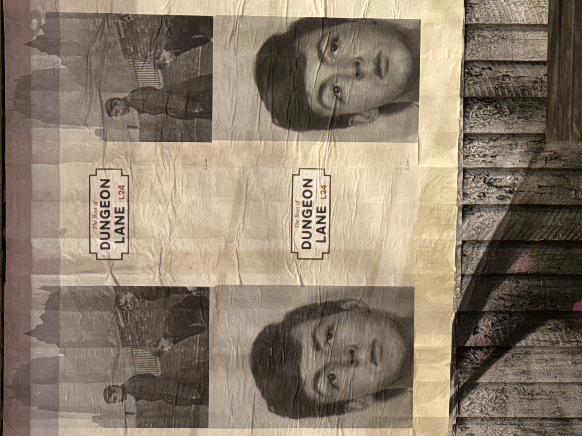 The image depicts a section of a public display, possibly street art, featuring photocopied posters affixed to a wall or surface. The posters contain two repeating images: a black and white photograph of a young boy's face, and a scene of two men in period attire standing near a building. Each poster also includes the text "The Boys of Dungeon Lane L24." The posters are affixed side by side. The background texture is rough and may depict wood or stone. The location, Dungeon Lane, suggests the display is located in the L24 postcode area of Liverpool, UK. The image has wrinkles and creases, indicating it's been pasted up and weathered. The posters are attached to the wall with tape.