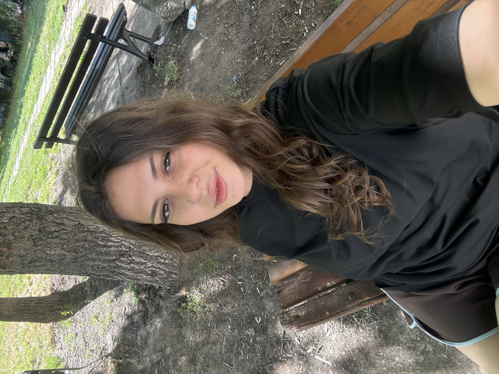 A young woman with long, wavy brown hair is taking a selfie while sitting on a wooden bench in an outdoor park. She is wearing a black t-shirt and brown shorts with light blue trim. The background shows trees, grass, and another park bench. The ground is covered in dirt and scattered leaves. The location is a park in Plovdiv, Bulgaria.