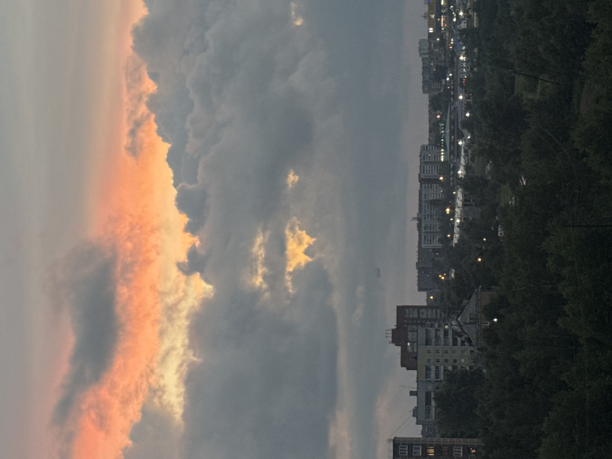 This image depicts a twilight view of a city, likely Bratsk, Russia, with a dramatic sky. The sky is filled with clouds, with the upper portion illuminated by an orange glow, suggesting a sunset or sunrise. Darker, grey clouds dominate the lower sky, with patches of light breaking through in some areas.
Below the sky, a cityscape is visible. Several multi-story buildings, characteristic of Soviet-era architecture, are prominent. In the foreground, a dense canopy of dark green trees obscures much of the ground level. Small lights are visible in some of the buildings and scattered throughout the treeline, indicating the presence of residential or commercial areas. A faint airplane is visible in the distance, flying high in the sky. The overall impression is of an urban environment at dusk, with natural elements of trees and sky interacting with man-made structures.