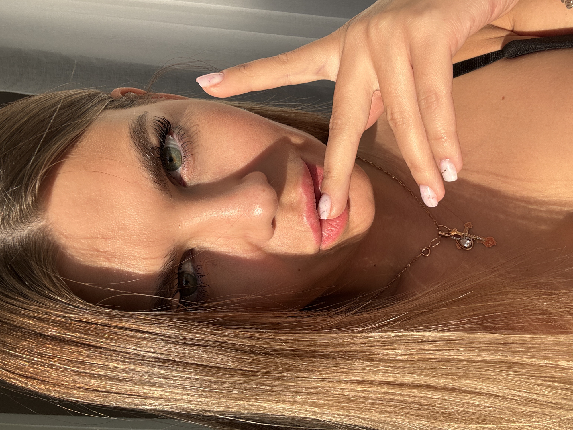 A close-up, eye-level, head and shoulders shot shows a fair-skinned woman with long, blonde hair. She is looking directly at the camera with green eyes and full, pink lips. Her right index finger is lightly touching her lips. She is wearing a black strap visible on her right side, and a gold chain with a cross pendant around her neck. The lighting suggests an indoor setting with natural light casting shadows across her face and hair. The background is out of focus, showing a sheer curtain or blind. There is no visible indication of the city of Sarapul, Russia, in the image.