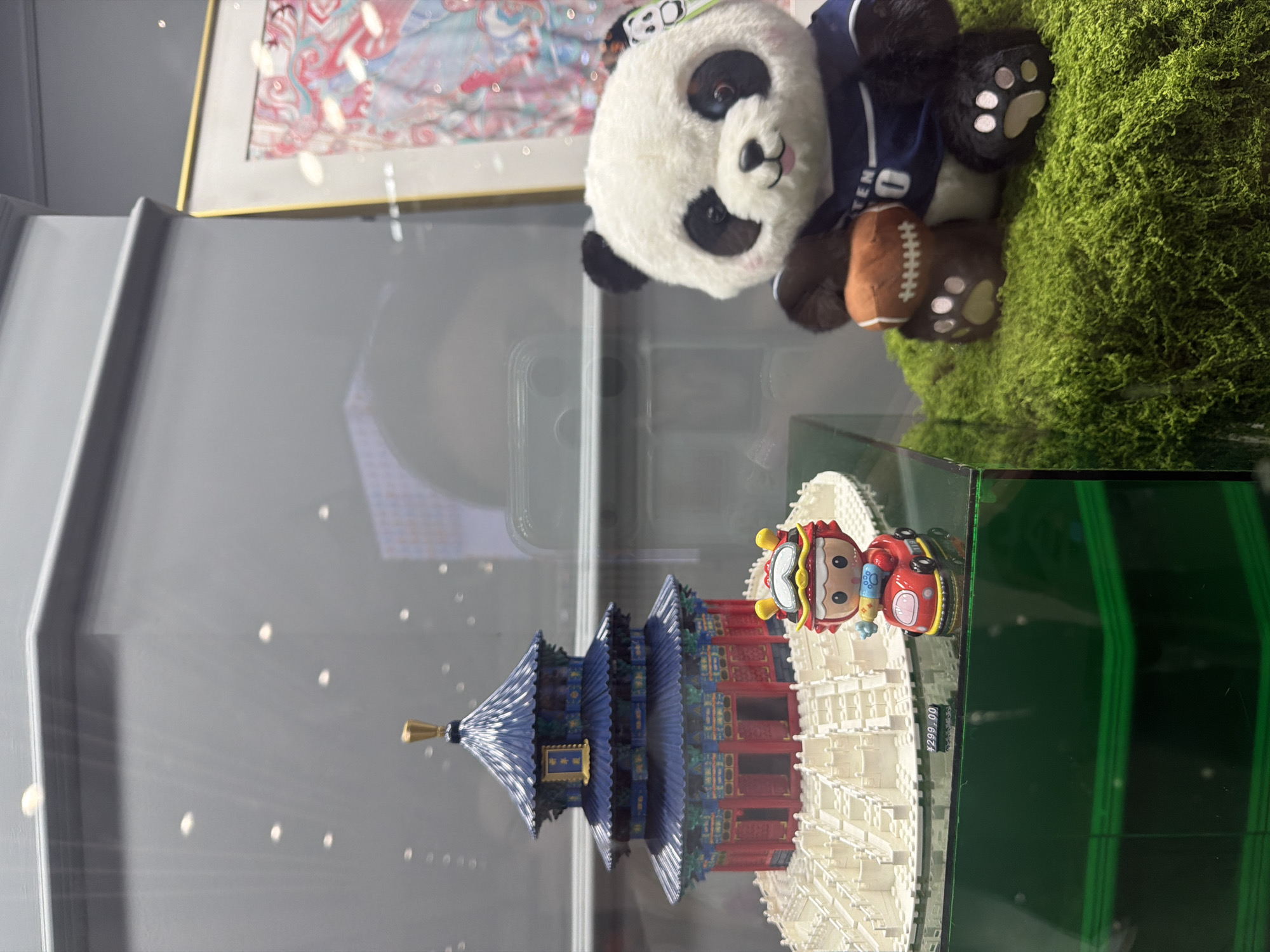 The image shows a display case in Chaoyang District, China, featuring two prominent items: a miniature replica of the Temple of Heaven and a plush panda toy. The Temple of Heaven replica, constructed from white blocks, has a blue tiered roof and red columns. A small, colorful character figurine on a red vehicle is positioned in front of the temple. To the right, a plush panda sits on a bed of artificial moss. The panda is wearing a blue shirt with white lettering and holds a brown football. A framed artwork with pink and blue patterns is visible in the background, reflected in the glass of the display case, along with other blurred reflections of people and objects. A price tag reading "¥299.00" is attached to the display base near the character figurine.