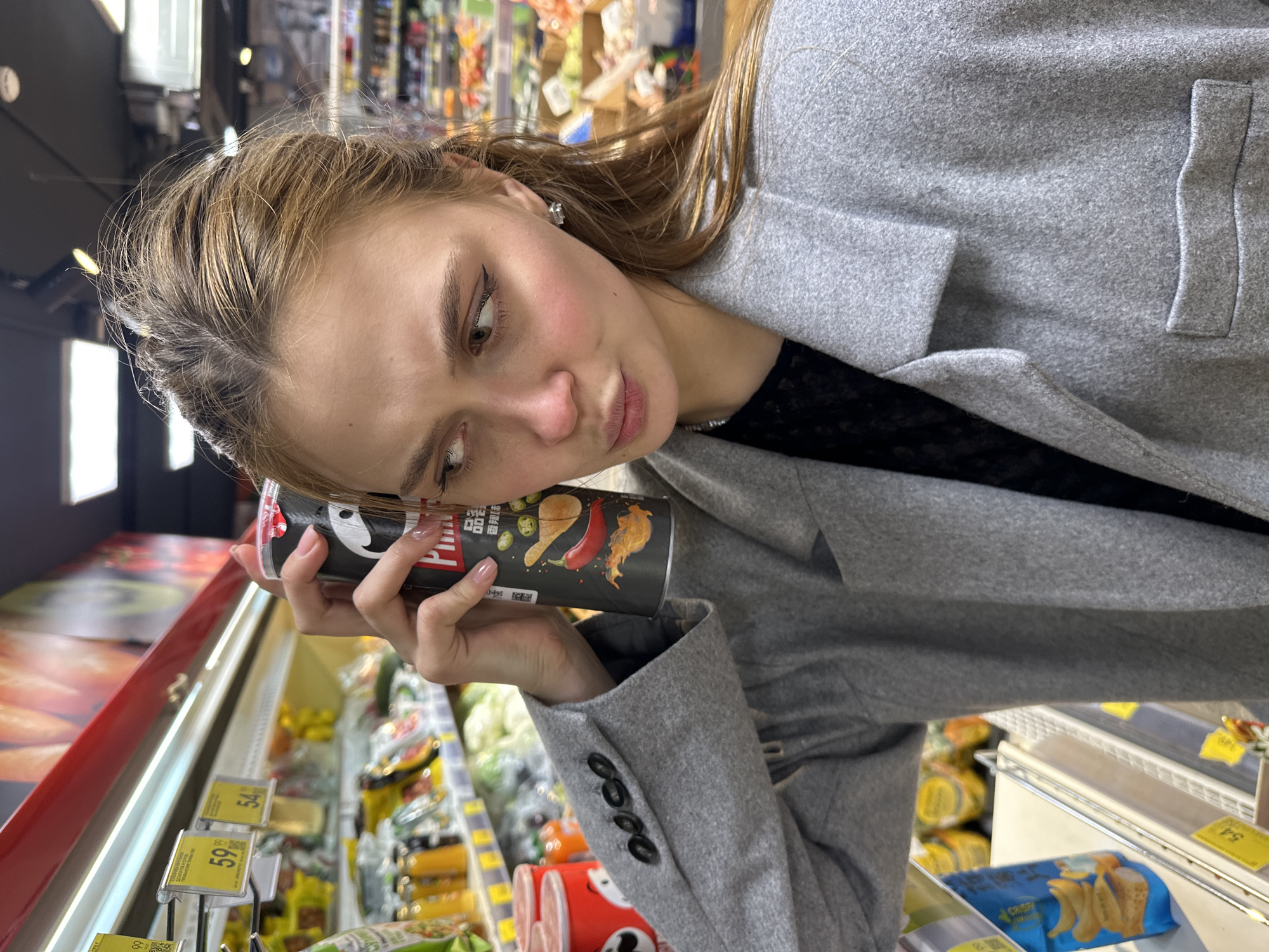 A woman stands in a grocery store in Krasnoyarsk, Russia. She is holding a can of Pringles potato chips against her face. The grocery store has produce and other food items for sale. Price tags are visible. The woman is wearing a grey coat over a black top.