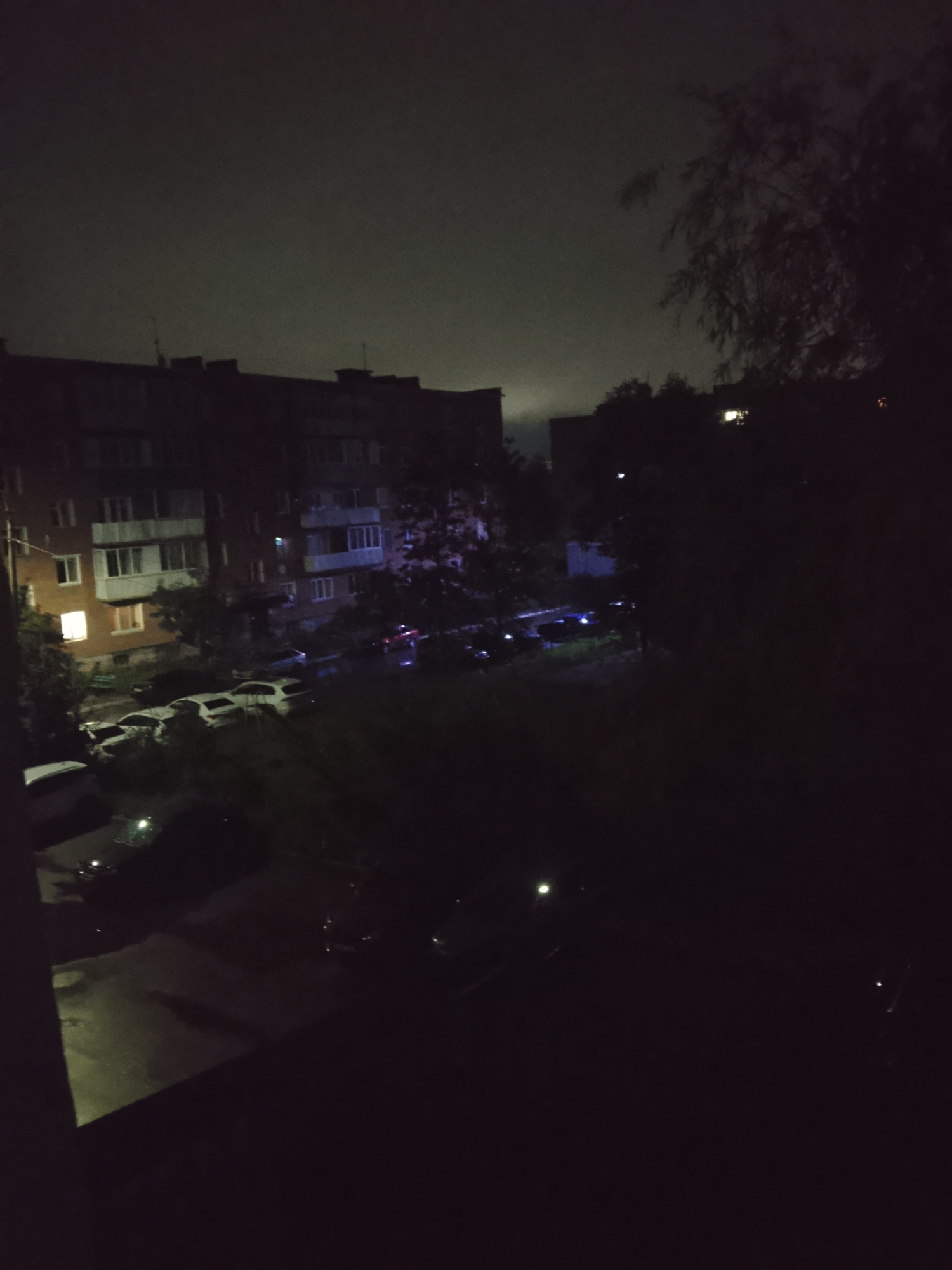 The image shows an exterior view of an apartment building and a street at night, possibly in Glazov, Russia. The building appears to be several stories tall with visible windows, some of which are illuminated. Several cars are parked on the street and in front of the building, and some have their headlights or taillights illuminated, suggesting they are either parked or in motion. The sky is dark, indicating nighttime, and there is some light pollution visible. Trees and foliage are present in the foreground and around the building, partially obscuring the view. The ground appears to be wet, possibly from rain, as indicated by reflections. There are no visible people in the image.