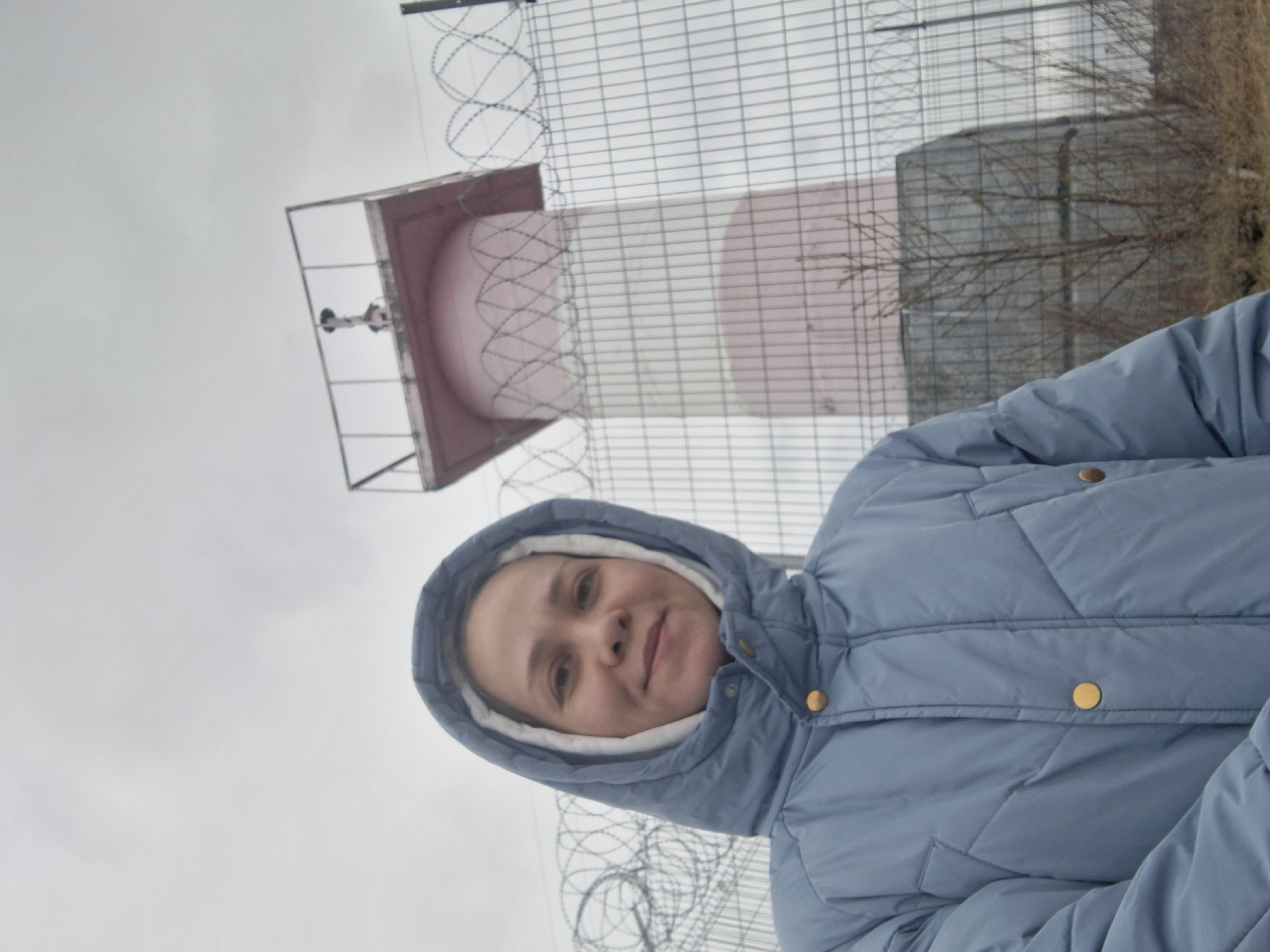 The image shows a person in a blue hooded jacket standing in front of a structure and fencing. The structure in the background is a striped tower, possibly part of an industrial facility or observation point. There is fencing with barbed wire visible. The sky is overcast. The person is wearing a hooded jacket with gold buttons. Based on the presence of a structure resembling a potential industrial landmark and the appearance of the fencing, the location may indeed be in the city of Chaikovsky, Russia, as per the prompt. However, without further information or specific landmarks, this is an educated guess.