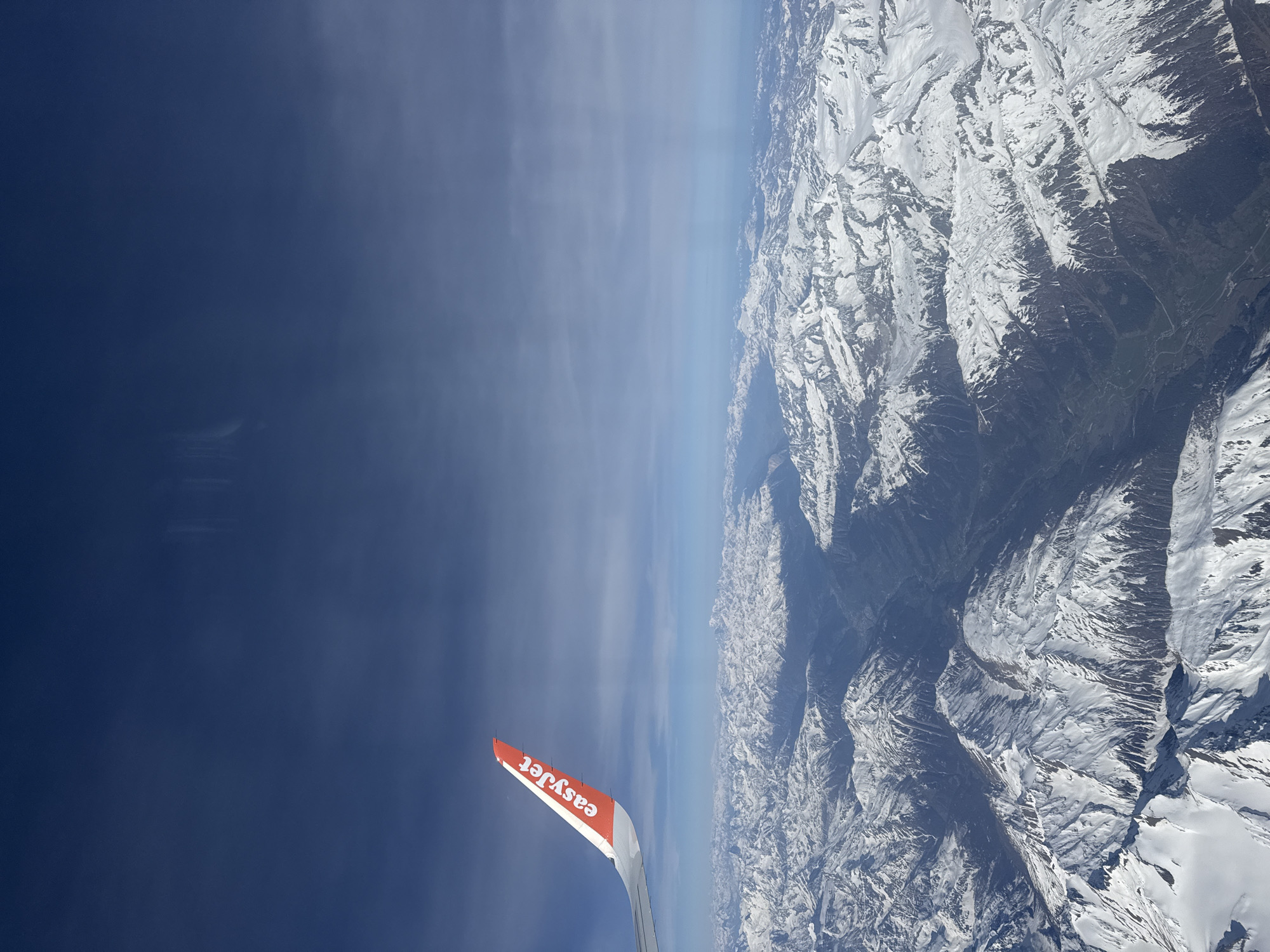 The image depicts a view from an airplane window, showcasing an EasyJet wing against a backdrop of snow-covered mountains and a clear blue sky. The EasyJet logo is visible on the wing. The mountains are rugged and heavily coated with snow. The sky is a deep blue, with some thin, wispy clouds near the horizon. The location appears to be a mountainous region, possibly the Alps or another similar range, but the exact city and country are unknown.