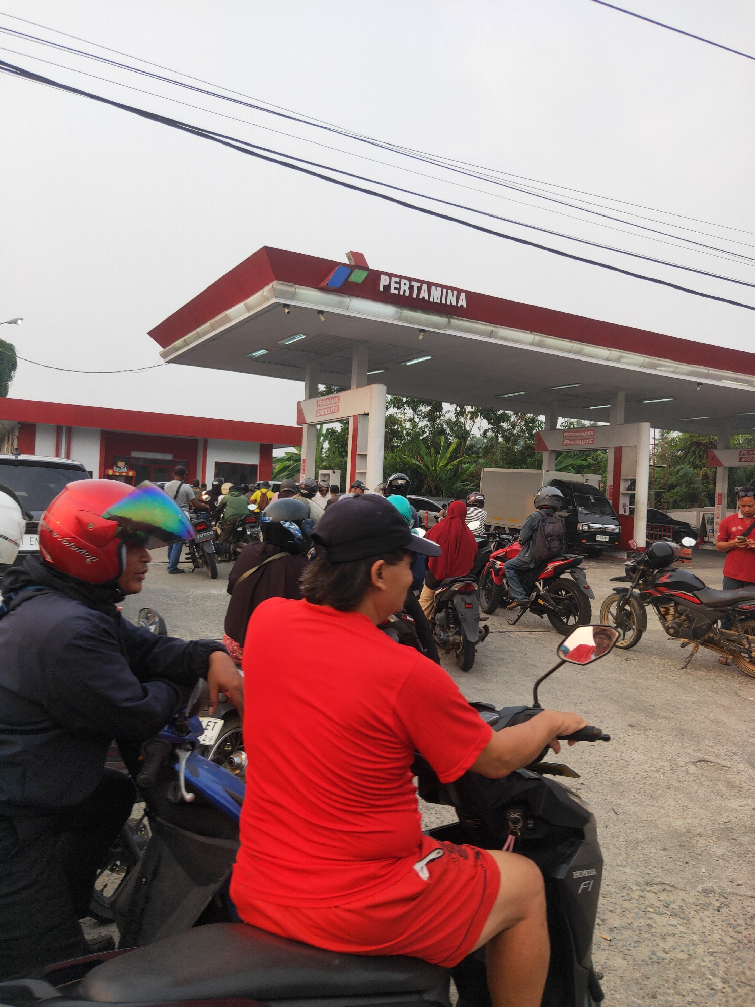 In Sintang, Indonesia, a street scene captures a line of motorcycles and a car waiting at a Pertamina gas station under an overcast sky. The station's red and white canopy, emblazoned with the Pertamina logo, is prominent. Several individuals are on or near their motorcycles, some wearing helmets, suggesting they are fueling or waiting to fuel. The ground is paved with a rough, grey surface. The atmosphere appears to be that of a typical day at a busy gas station.