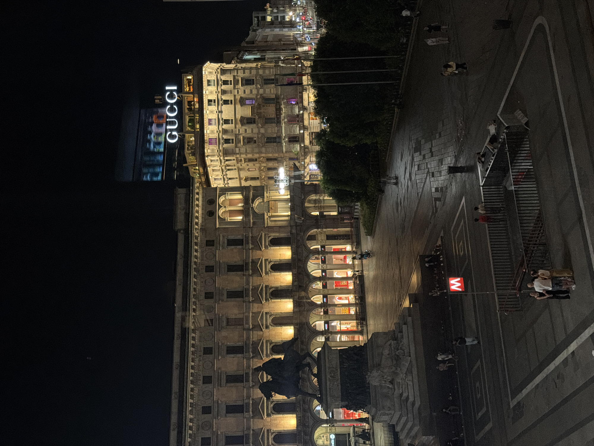 This is an aerial nighttime photograph of Piazza del Duomo in Milan, Italy. The scene captures the iconic Duomo and Galleria Vittorio Emanuele II, illuminated by artificial lights. In the foreground, a statue of a man on horseback stands on a pedestal. To the right, a building with a large "GUCCI" sign is visible. People are seen walking and gathered in the square, with some sitting on benches. A red "M" sign, indicating a metro station, is also present. The square is paved with cobblestones, and trees and streetlights line the perimeter. The overall atmosphere is lively, with evidence of urban nightlife.