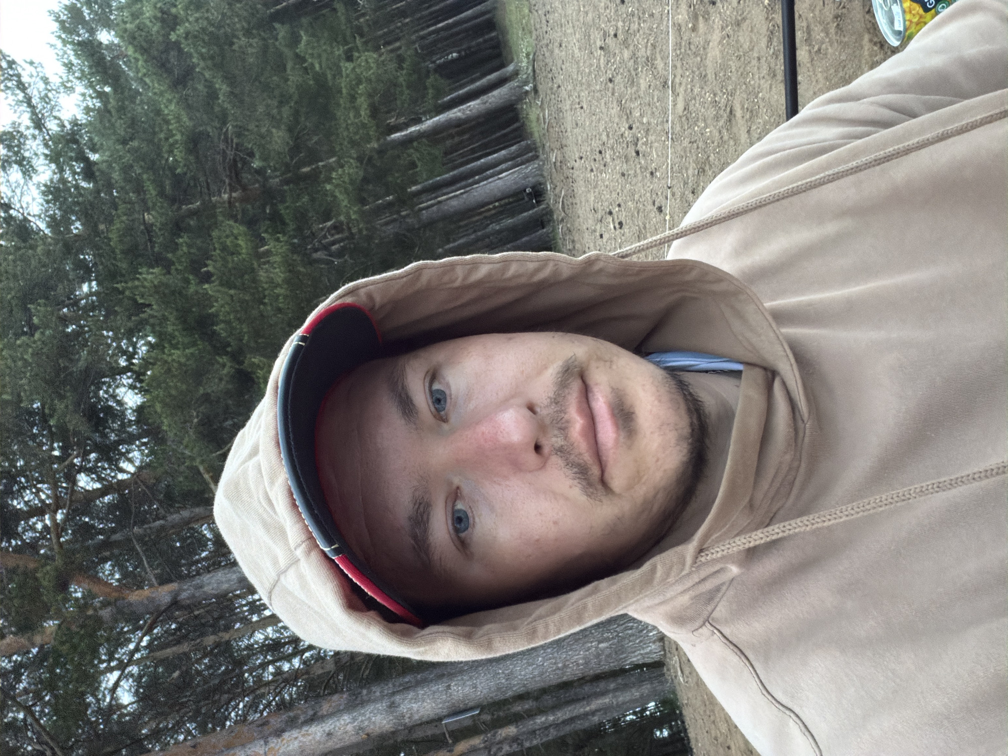 A forensic description of the scene, located in the city of Chaikovsky, Russia, reveals an adult male positioned centrally in an outdoor environment. The individual is looking directly into the camera with a neutral expression. He has light skin, blue eyes, and light brown hair partially visible under a red and black baseball cap, which is in turn covered by the hood of a light brown/beige pullover hoodie. A light blue garment collar is visible beneath the hoodie. The hoodie features visible drawstrings at the neck.
The background consists of a dense coniferous forest dominated by tall pine trees, their trunks stretching vertically. The ground is dry, light brown soil or sand, with sparse debris such as pine needles or small cones. The lighting is soft and diffused, indicating an overcast sky. To the lower right of the subject, partially visible on the ground, are a yellow and green can or container, a black horizontal bar or pole, and a thin white line or rope extending horizontally across the frame.