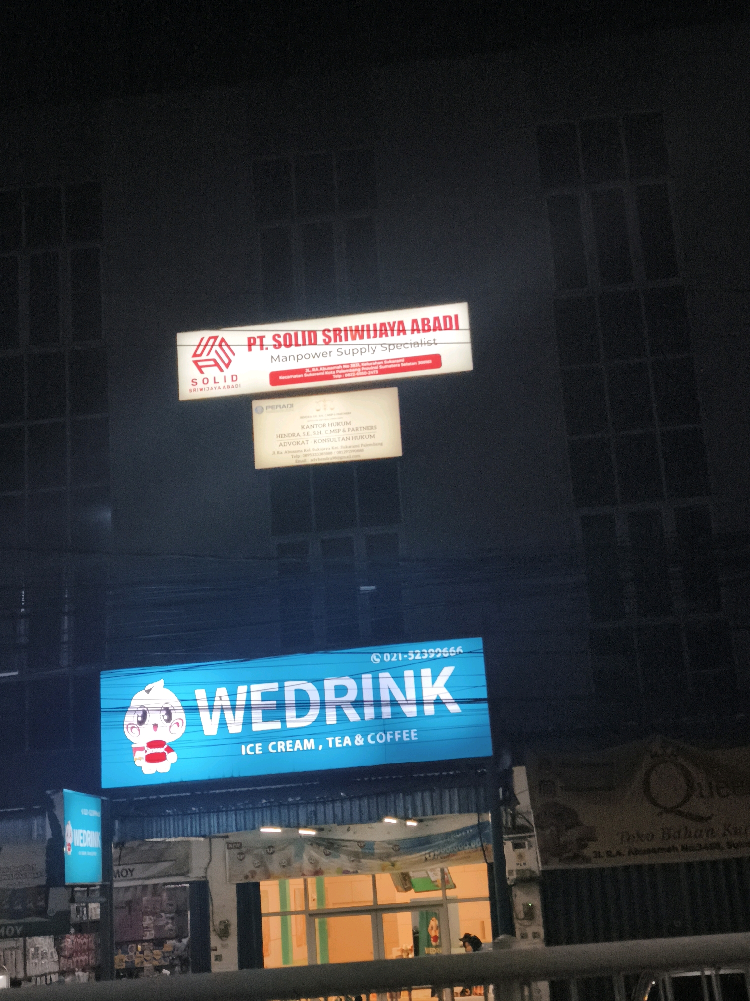 This is a nighttime, exterior shot of a commercial building in Palembang, Indonesia. Prominently displayed are three illuminated signs. The topmost sign, a horizontal rectangle with a red and white logo, reads "PT. SOLID SRIWIJAYA ABADI Manpower Supply Specialist." Below this, a smaller, cream-colored rectangular sign with a black logo displays "PERADI KANTOR HUKUM HENDRA S.H., C.M.S.P & PARTNERS ADVOKAT KONSULTAN HUKUM." The address is listed as "Jl. RA. Abusamah No. 2835, Kelurahan Seberang Ulu Ilir, Kecamatan Seberang Ulu Ilir, Palembang." A third, large, bright blue horizontal sign features a cartoon character resembling a steamed bun and the text "WEDRINK ICE CREAM, TEA & COFFEE." A smaller, vertical blue sign with "WEDRINK" is also visible to the left of the larger sign. A banner with "MOY" is visible below the "WEDRINK" signs. The background consists of dark, silhouetted building facades with window grids. The overall impression is that of a commercial street scene at night.