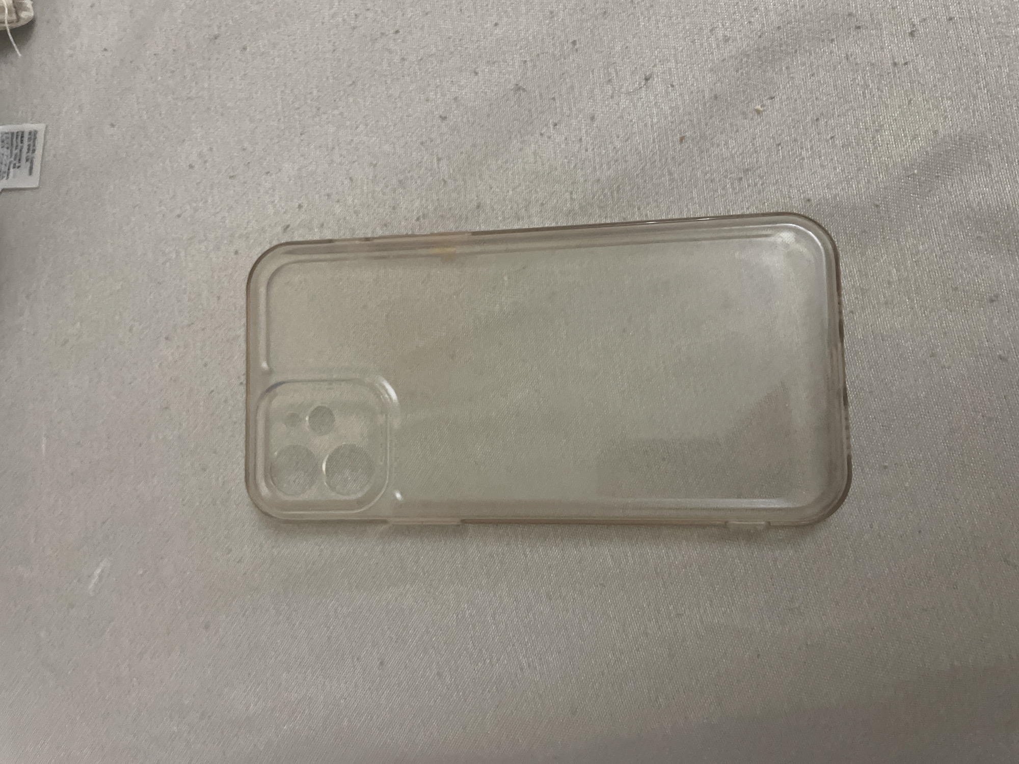 The image displays a transparent silicone phone case, exhibiting a slight yellow discoloration consistent with wear, particularly along its edges. The case is designed for a smartphone model featuring a triple-lens camera system, indicated by the three circular cutouts in the upper left corner of the case. It rests centered on a light-colored, textured fabric surface, likely a textile such as cotton or linen, which shows minor pilling and faint smudges. A small, white fabric tag with unreadable text is partially visible in the top right portion of the background. The scene is situated in the city of Ihtiman, Bulgaria. No individuals, actions, or interactions are present.