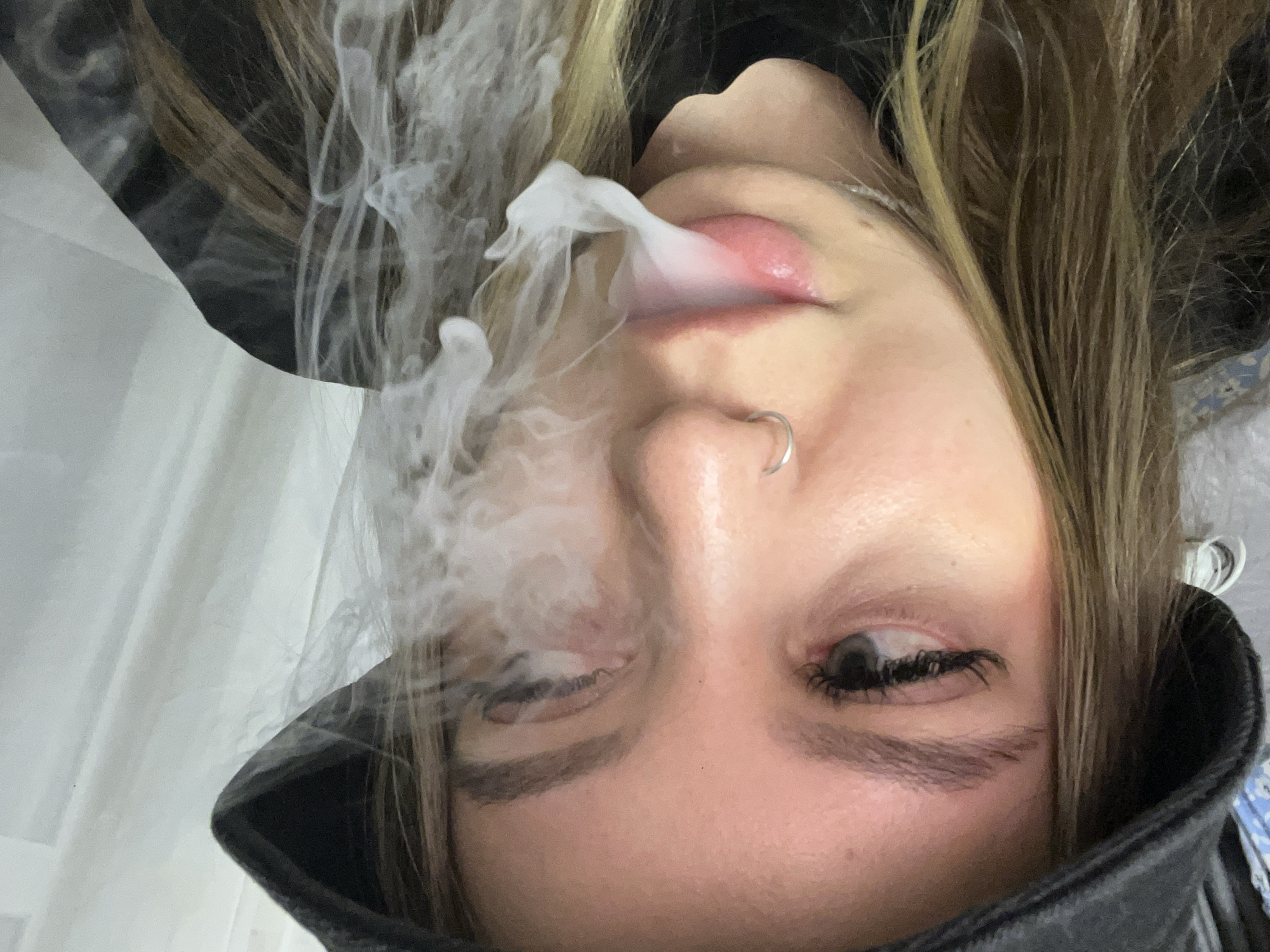 A close-up, inverted portrait captures a person, likely female, exhaling a dense cloud of white smoke or vapor from their mouth. The individual has fair skin, light brown or blonde hair, and a silver hoop nose piercing on the right nostril. Prominent dark eyelashes frame their eyes; the left eye is partially obscured by the smoke, while the right eye looks downwards and to the side. Their lips are slightly parted, appearing glossy.
The person wears a dark-colored garment, possibly a jacket or hoodie, with a collar visible around the chin. The background on the left features a soft, blurred white or light-colored surface, resembling a curtain or wall. A small segment of patterned blue and white fabric is visible on the far right. The image focuses tightly on the face and the exhaled smoke, indicating an activity of smoking or vaping. No specific time of day or weather is discernible, and no text is visible. The scene takes place in an indoor setting in Krasnodar, Russia.
