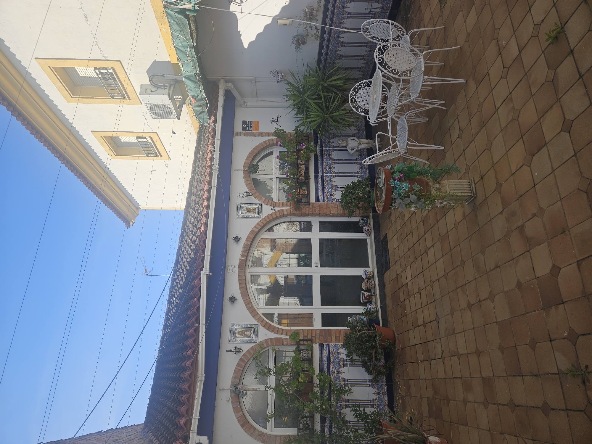 The image depicts an outdoor courtyard in Pedro Abad, Spain. The courtyard is paved with brown tile bricks. A building with arched windows and decorative tiles borders one side of the courtyard. White metal tables and chairs are arranged on the tiled surface. Potted plants and flowers are scattered around the courtyard, adding greenery to the space. An adjacent building is visible on the opposite side. The sky above is clear and blue.