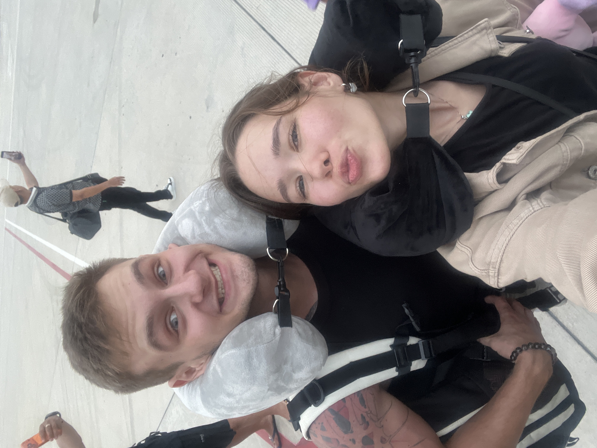 A bright and lively selfie captures a cheerful couple outdoors, likely at an airport or a travel hub in Bac Cam Ranh Ward, Vietnam. The image is taken from a slightly high angle, looking down.
In the foreground, a man with short brown hair and blue eyes smiles broadly at the camera. He wears a black t-shirt, a grey neck pillow, and a black and white harness system, possibly part of a parachute rig or a substantial backpack. A beaded bracelet adorns his right wrist, and tattoos are visible on his right arm. He holds an orange object, possibly a phone or a remote, in his left hand.
Resting her head on the man's right shoulder, a woman with brown hair and light features makes a playful "kiss face" or pout towards the camera. She wears a black top, a beige jacket, and a black neck pillow. A delicate necklace with a light-colored pendant is visible around her neck, and she wears a small earring.
The background shows a large paved area with faint red and white markings on the ground, suggesting an outdoor space like an airport tarmac. Several other individuals are visible in the distance; one person wearing a light hat and patterned shirt is holding a phone up, seemingly taking a photo or video. The scene is brightly lit, indicating daytime. The couple appears happy and relaxed, suggesting a pleasant travel experience or activity. No visible text is clearly discernible.