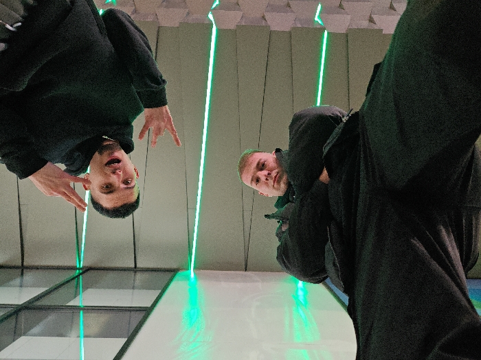 The image depicts two young adult males in a modern indoor setting within Shumen, Bulgaria, presented from a low-angle perspective which renders them visually inverted. The male on the left, with short dark hair and a light beard, wears a black hooded sweatshirt and a silver chain. He smiles openly towards the camera, extending his left hand to form a 'V' sign and his right hand in a gesture with extended index and pinky fingers. The male on the right, partially visible, has short light-colored hair and wears a dark jacket and trousers, maintaining a neutral gaze towards the camera. The background consists of a light-toned ceiling structure featuring multiple parallel, linear green LED light strips. Below the subjects, a reflective, light-colored floor or platform is visible, showing illuminated streaks from the green lighting. The environment is characteristic of a contemporary public space or commercial complex.