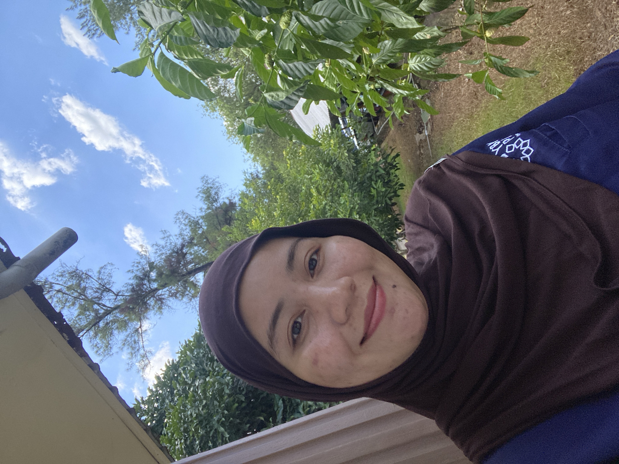 A woman wearing a brown hijab and a blue shirt takes a selfie outdoors. She is smiling and looking at the camera. Behind her, there are green trees, a clear blue sky with white clouds, and a roof of a building. The scene appears to be in a residential area or a garden. The image is taken during the day.
