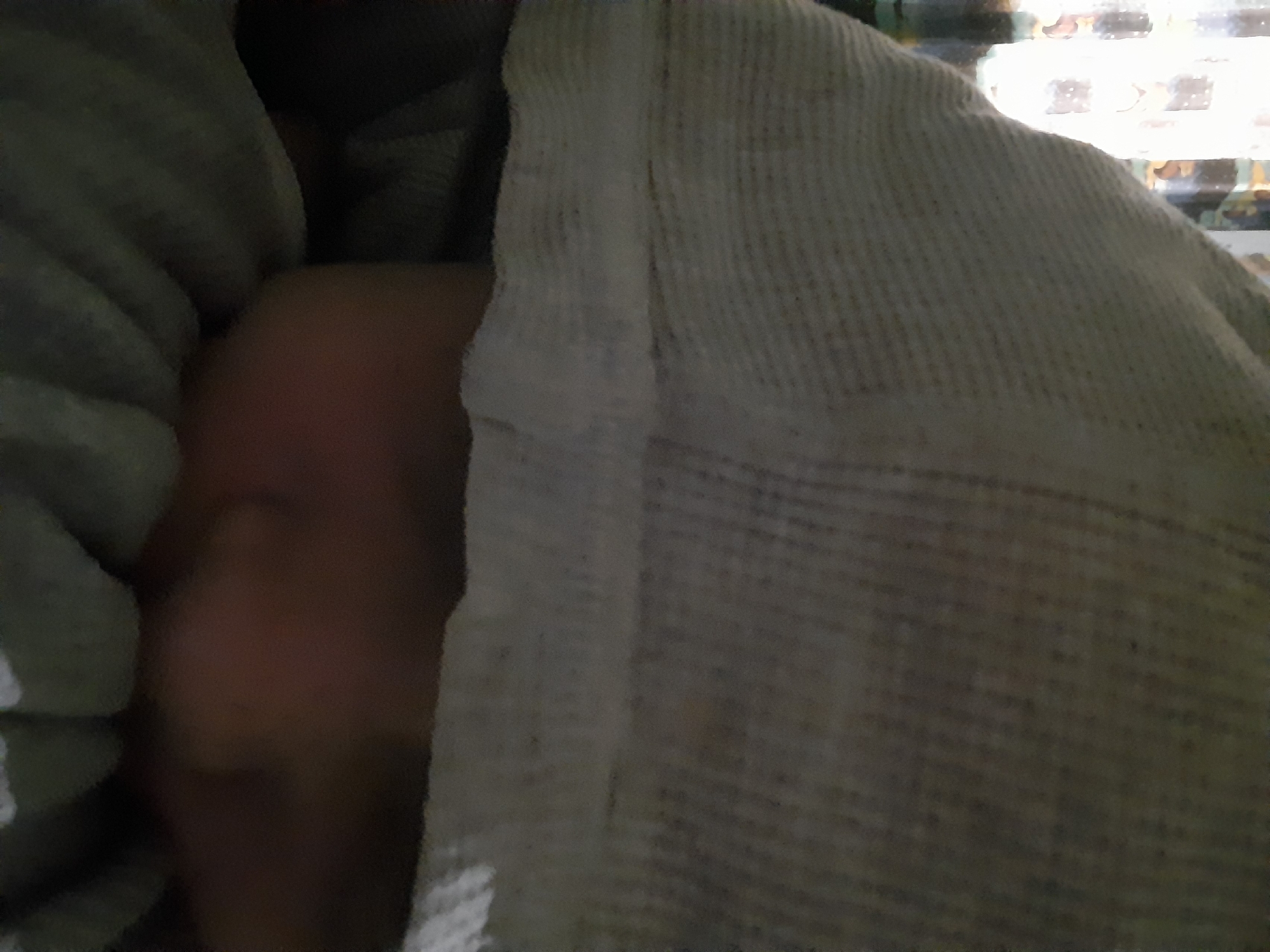 The image shows a close-up, low-angle shot of a person, likely an infant, wrapped in blankets. The person's face is partially visible, with one cheek and eye appearing to be in shadow. The blankets are textured, with a waffle-knit pattern visible on the white blanket in the foreground and a ridged texture on the olive-green blanket on the left. The lighting is dim, suggesting an indoor setting, possibly at night or in a dimly lit room. There are no clear indications of the weather, time of day, or specific location beyond the general context of Santa Ana, Venezuela. The overall mood is one of quiet rest or sleep. There is no visible text in the image.