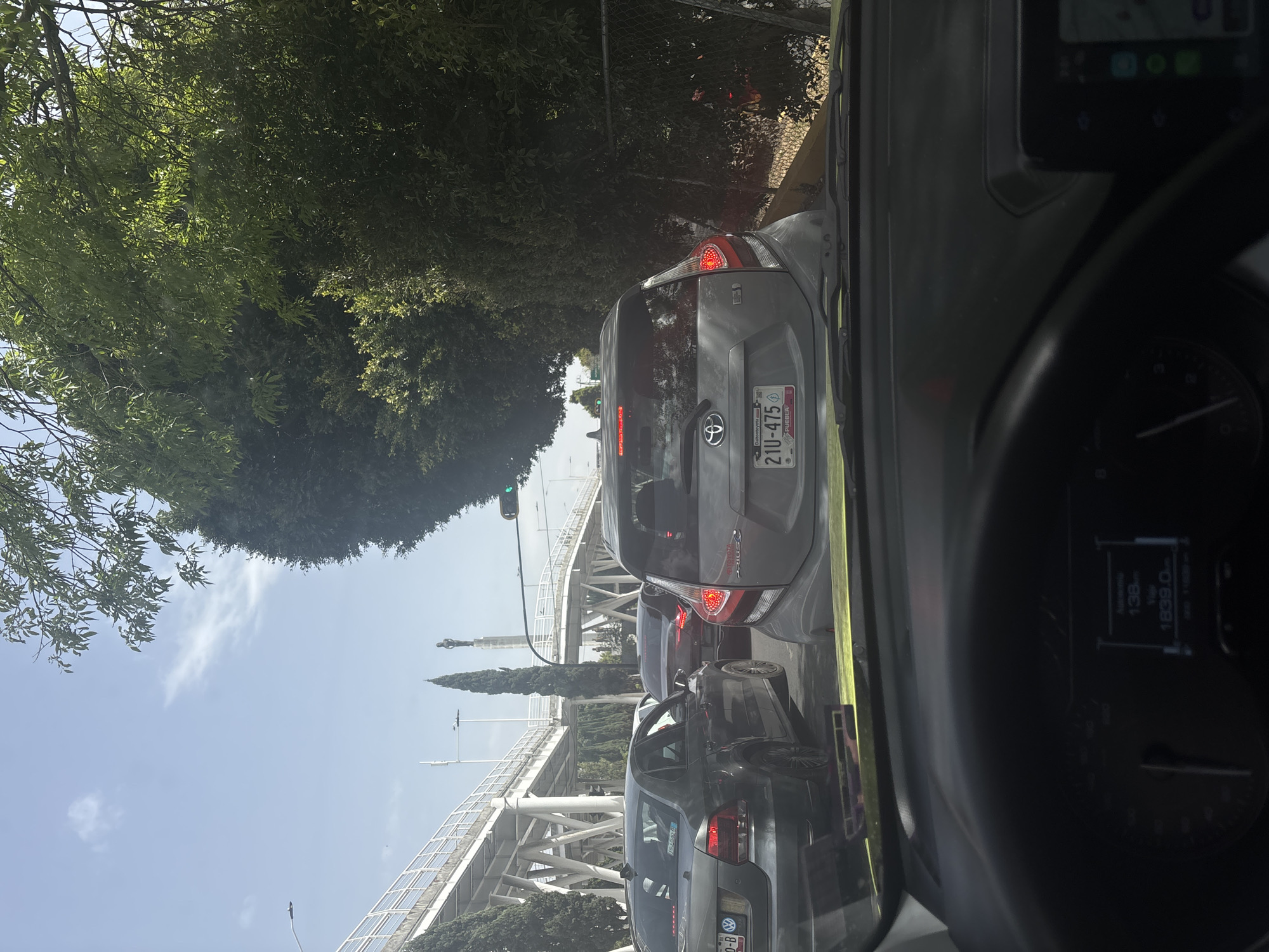 The image shows a congested street scene in Puebla, Mexico, viewed from inside a car. The license plate on the car directly ahead reads "21U-475" with a "PUEBLA" designation, suggesting the location is in the state of Puebla. There are other cars in front and behind the camera car, indicating a traffic jam. A large pedestrian bridge is visible in the distance, potentially a recognizable landmark within the city. Trees and greenery line the side of the road. The sky is blue and partly cloudy. The car's dashboard and steering wheel are partially visible in the foreground, including a digital display with mileage information.