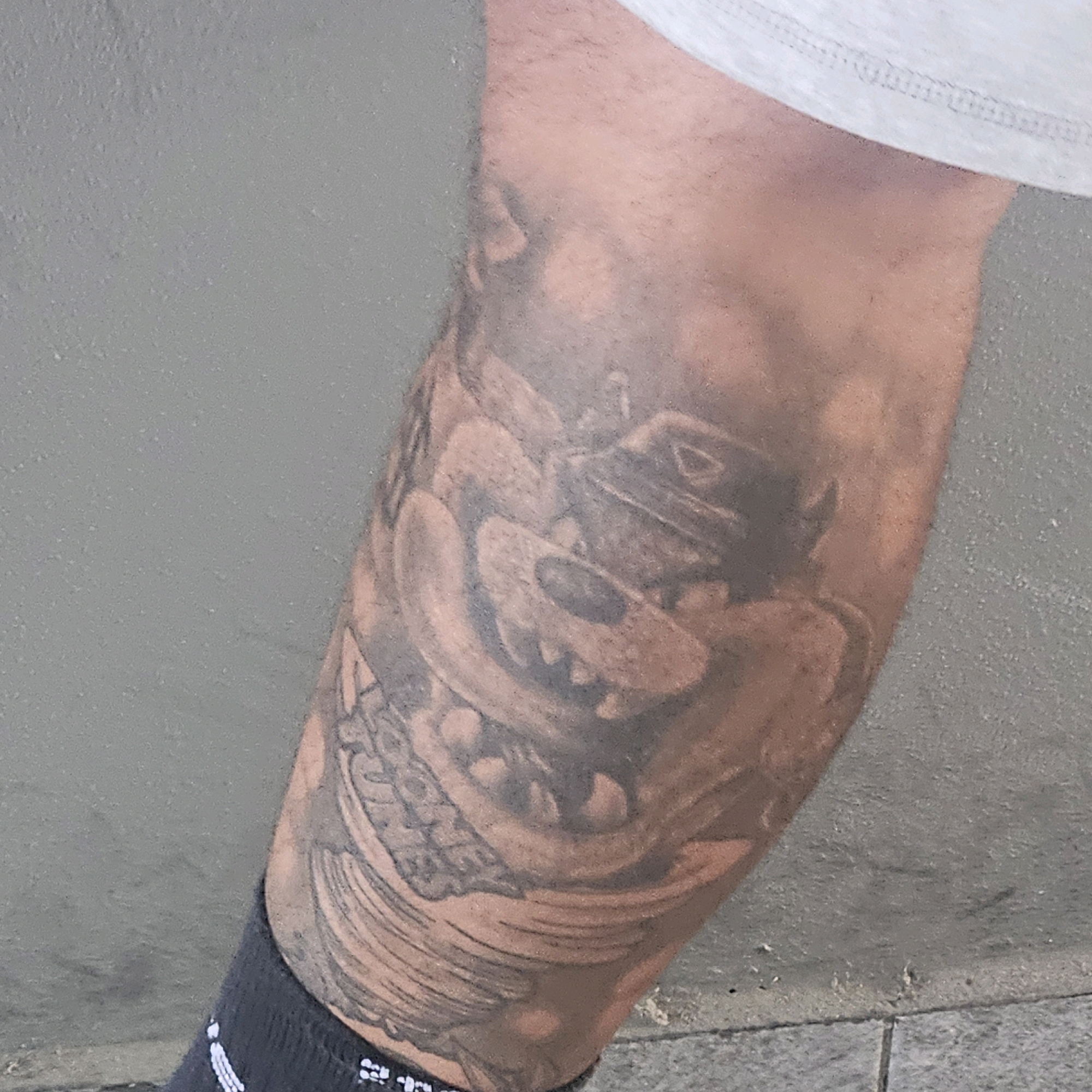 An incomplete tattoo of a cartoon character, possibly Bugs Bunny, is visible on a person's leg against a grey concrete wall. The tattoo is rendered in shades of grey and black and appears to be partially faded or in progress. The character's face, with large eyes and teeth, is the most discernible feature. The person is wearing a grey t-shirt and black socks. The background suggests an outdoor urban environment. The location is identified as Poirino, Italy.