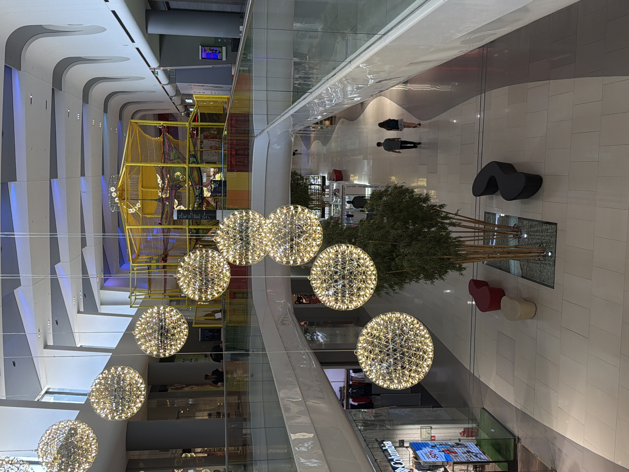 The image shows an interior view of a modern shopping mall. Suspended from the ceiling are several large, spherical chandeliers made of intricate metalwork and illuminated by numerous small lights. In the background, a brightly colored indoor playground structure with yellow netting is visible.
On the lower level, people are walking in a spacious, tiled corridor. To the left, a store with a glass facade displays clothing. To the right, a curved white railing overlooks the lower level. Further down the corridor, a couple is walking away from the camera.
On the lower level floor, there are several seating areas, including red, white, and black benches, and a cluster of bamboo plants.
The mall features a contemporary architectural design with curving white walls and a high ceiling. The lighting is bright, and the overall impression is one of spaciousness and modern design.
This scene is located in the OZ Mall in Krasnodar, Russia.