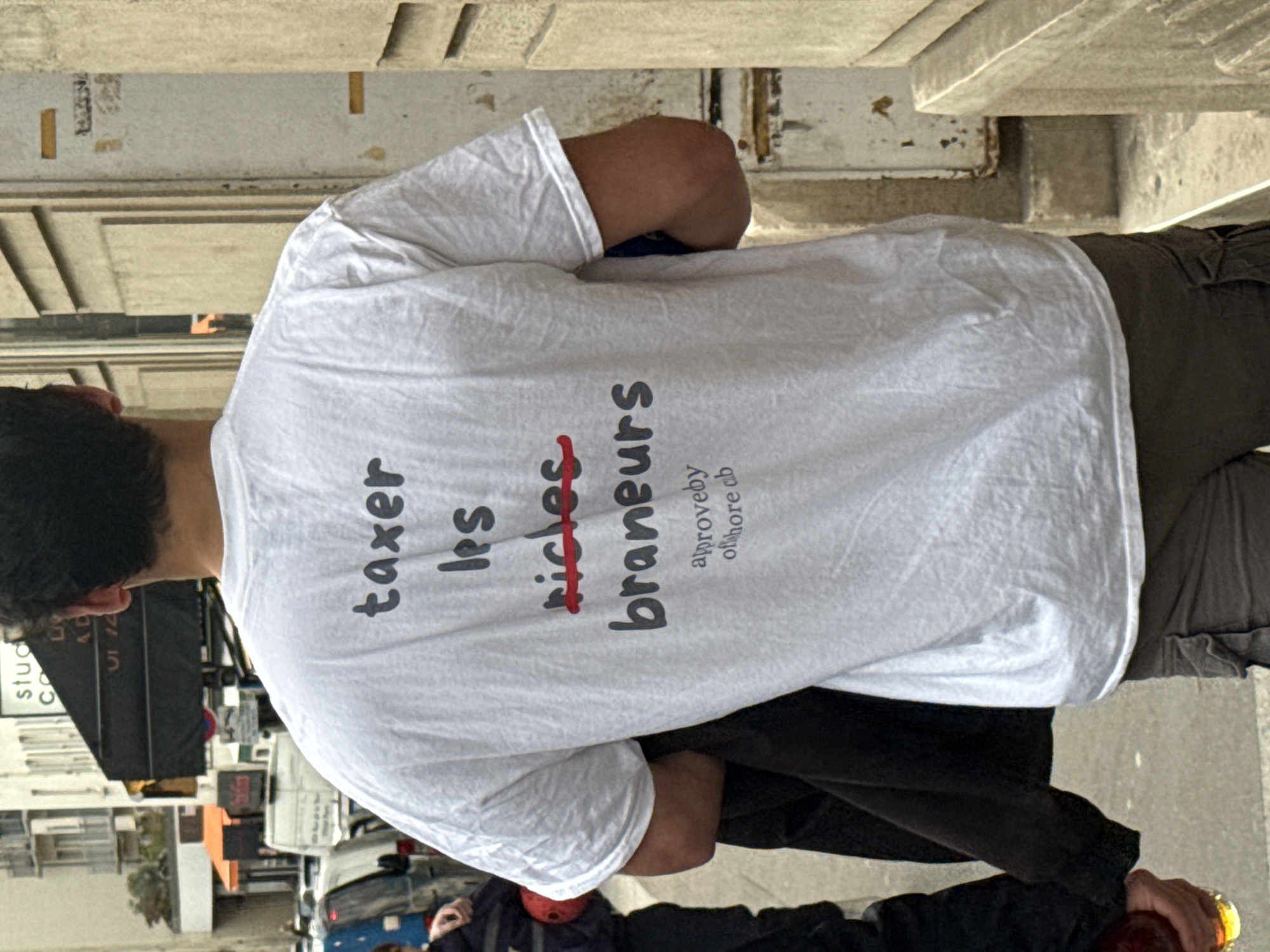 A man in tan trousers and a white t-shirt stands on a sidewalk. The back of his t-shirt has text printed in black that reads "taxer les" with the word "riches" (rich) in red with a line through it. Underneath the crossed-out "riches" is the word "braneurs." Below that, in smaller grey text, is "aprove by offshore cb." The man has dark hair and is wearing a black jacket that is draped over his arm. He is walking down a sidewalk. In the background are buildings and cars, with storefronts visible. This scene likely takes place in Paris, France, considering the French text on the shirt and the architectural style.
