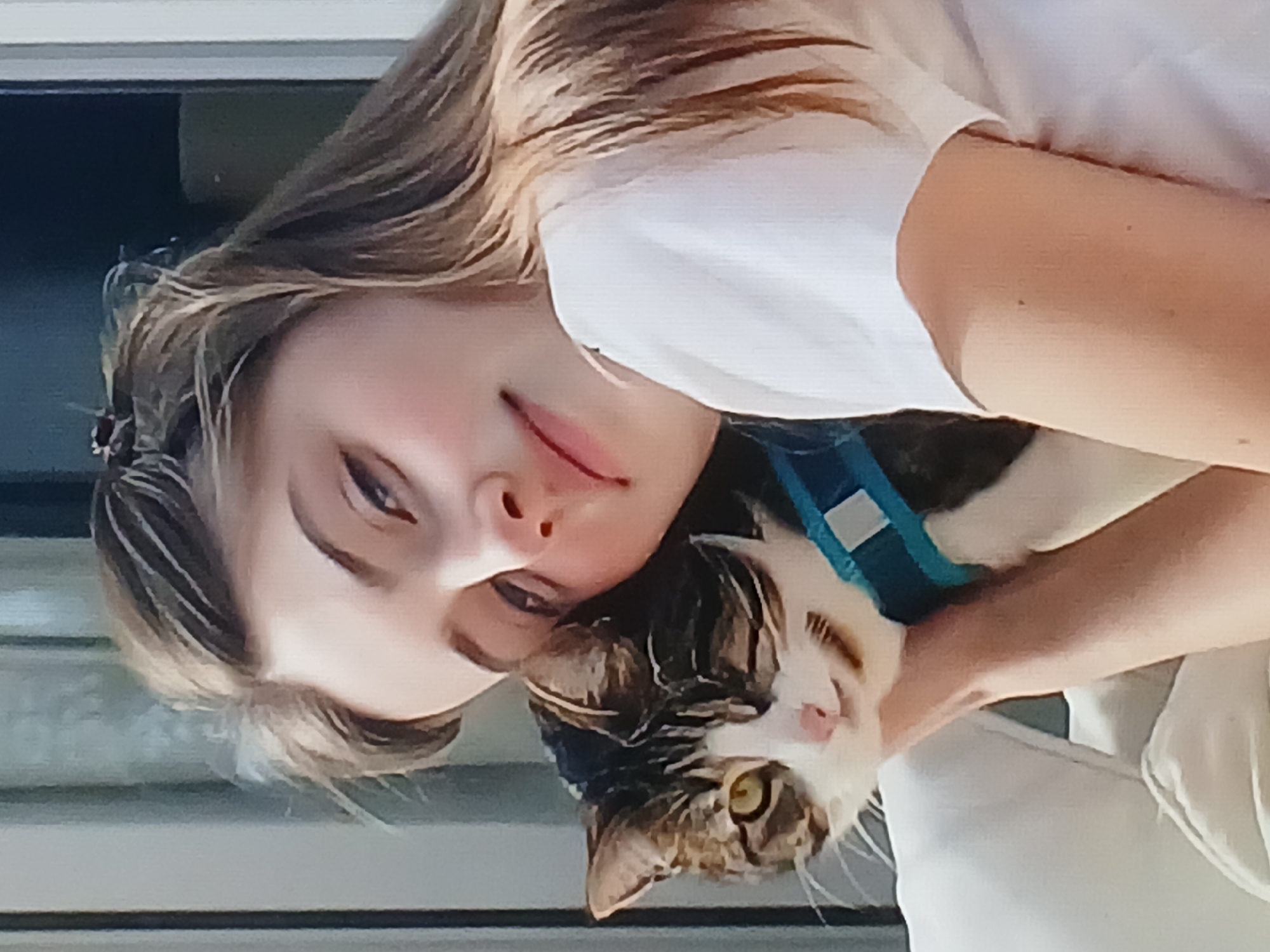 A close-up, waist-up shot shows a young person with light brown hair embracing a tabby cat. The person is wearing a white t-shirt and has their head tilted, with their cheek resting against the cat. The cat, facing the camera, has green eyes and is wearing a turquoise harness. They are both positioned on a light-colored surface, possibly a couch. The background is blurred, but appears to be an indoor setting with vertical blinds and a dark area. The image was captured in Dnipro, Ukraine.