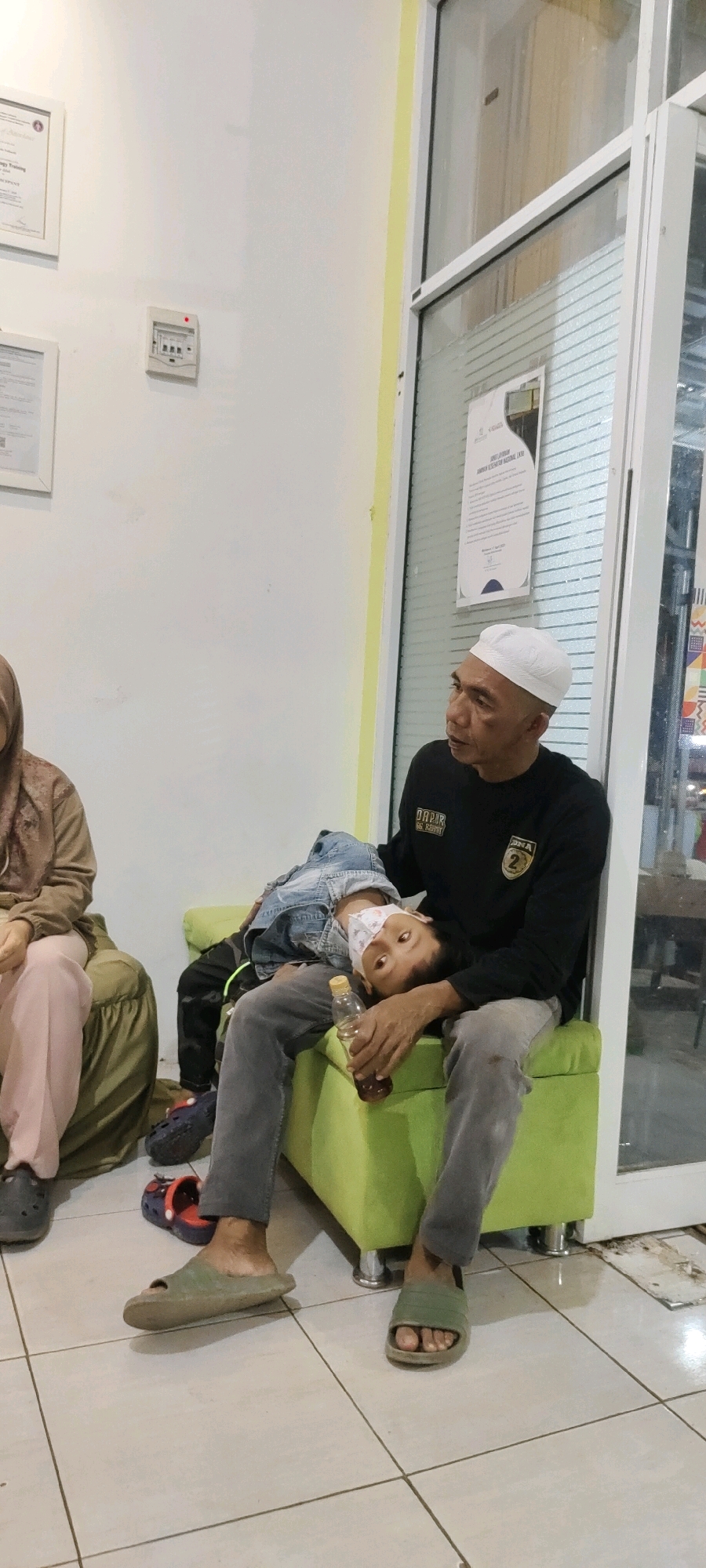 A man wearing a white skullcap and a black long-sleeved shirt sits on a lime green sofa in what appears to be a waiting room. He holds a young child, wearing a denim jacket and a face mask, in his lap. The child's head rests on the man's arm, and the child is looking upwards towards the man. A partially full bottle of liquid with a yellow cap is held by the man, resting on his leg. Another person, partially visible, sits on a green chair to the left of the sofa. The room has light-colored tiled flooring and a white wall with framed documents and an electrical panel. A large glass door with a poster on it is visible to the right of the sofa. The image was taken in Sungai Sipai, Indonesia.