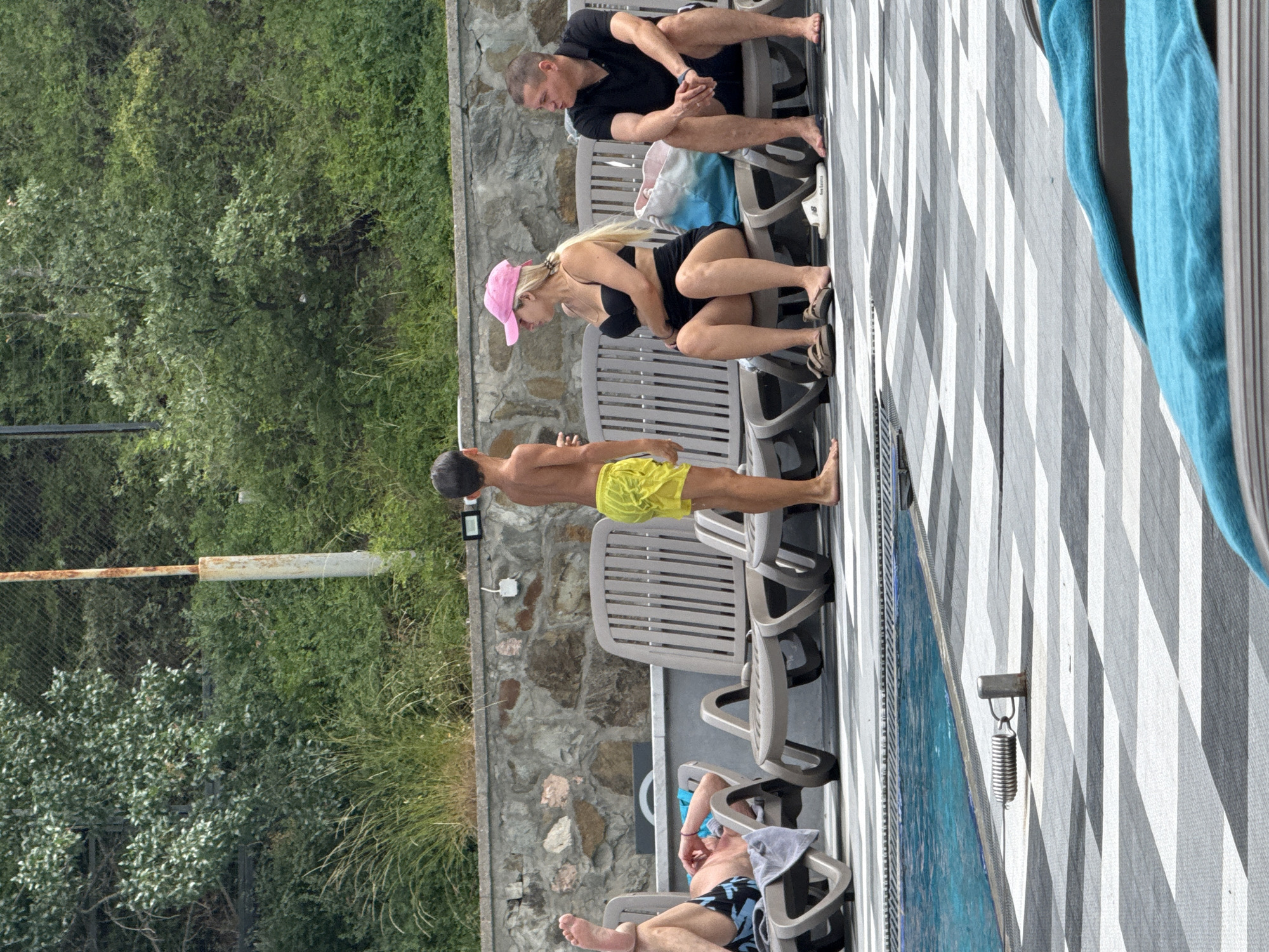 The image shows a group of people lounging by a pool. In the foreground, a man in yellow swim trunks is lying on a chair, facing away from the camera. Next to him, a woman in a black bikini and pink hat is also seated on a chair, looking down. Further back, a man in a black t-shirt and shorts is sitting on a chair, his arms crossed. The scene is set on a patio with a patterned floor and rows of empty chairs. In the background, there is lush green foliage and a stone wall. The overall atmosphere appears relaxed and leisurely, suggesting a sunny day at a resort. The presence of a swimming pool and lounge chairs indicates an outdoor recreational setting, likely in a warm climate. There are no discernible timestamps or weather cues, but the lighting suggests daytime. The location context of Yalta, Ukraine, places the scene in a coastal resort town.