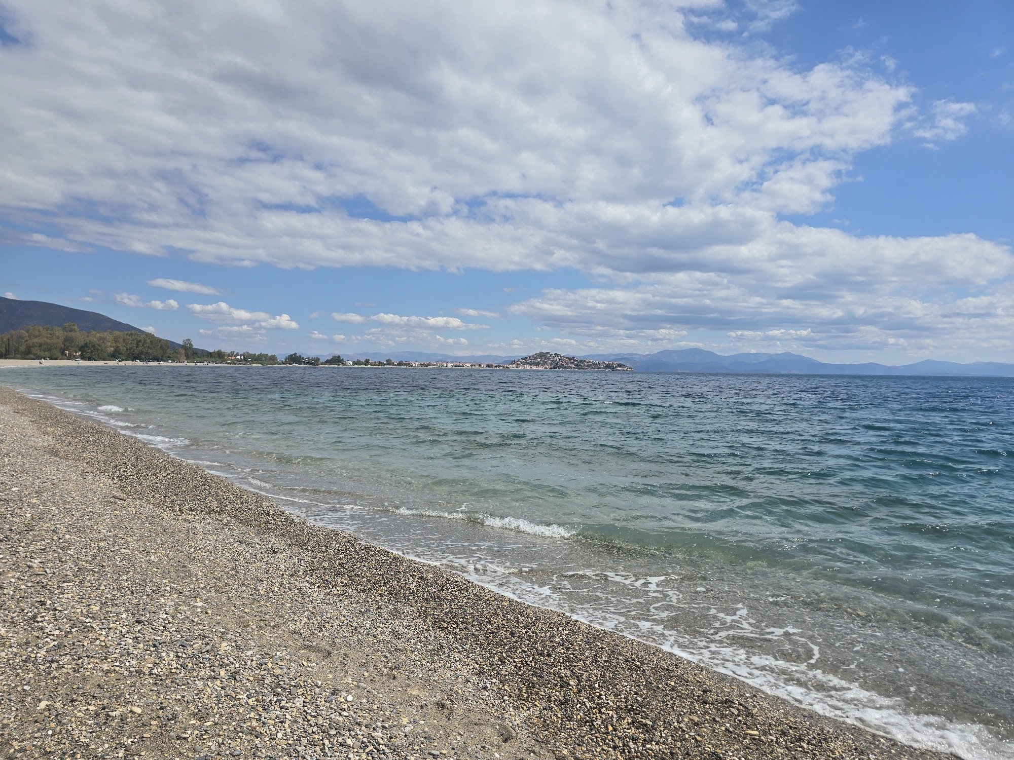 The image depicts a scenic view of a pebble beach in Astros, Greece. The foreground is dominated by the textured surface of the beach, covered with various sizes and colors of pebbles. Gentle waves lap at the shore, creating a thin line of white foam. The sea is a clear, turquoise hue, stretching towards the horizon. In the mid-ground, a distant coastline is visible, with a cluster of buildings perched on a hill, suggesting a small town or village. Beyond the coastline, a range of mountains forms a hazy silhouette against the sky. The sky itself is a vibrant blue, dotted with a mix of white, fluffy clouds and larger, more dispersed formations. The overall impression is one of a tranquil, natural setting with a touch of coastal habitation.