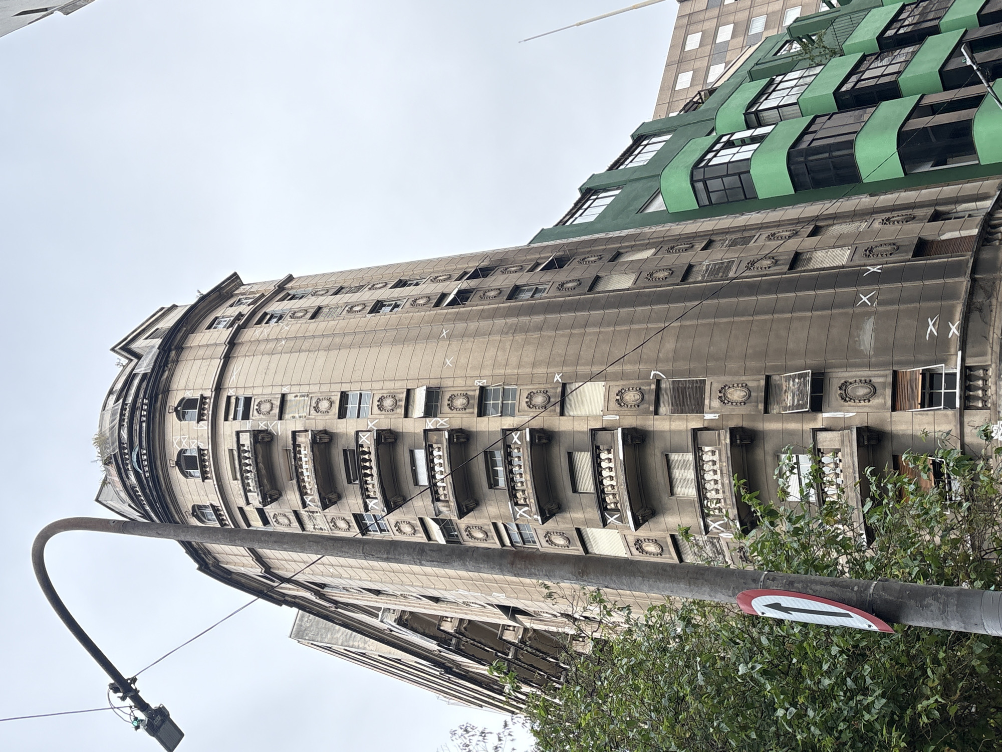The image shows a low-angle view of an old, curved, beige building with decorative balconies and windows. To the right of this building is a modern, green building with large glass windows. A tall, grey lamppost with a street light and a traffic sign is in the foreground, partially obscuring the beige building. Trees are visible to the left. The sky is overcast. This scene is located on Rua Augusta in the Consolação neighborhood of São Paulo, Brazil, specifically in front of the Edifício Planalto.