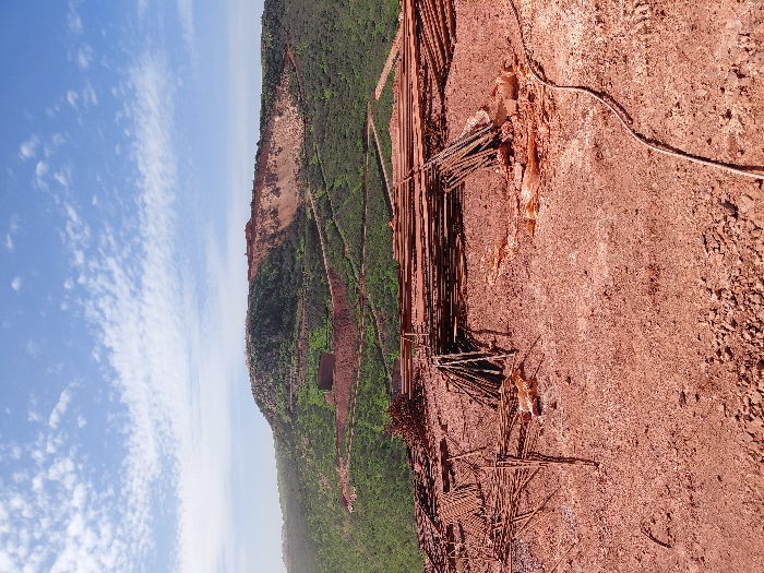 The image displays an outdoor scene under a blue sky with scattered white clouds, depicting a landscape characterized by industrial or mining activity. The foreground consists of uneven, reddish-brown earth mixed with small rocks and gravel. Prominently featured are multiple quantities of rusty metal rebar: a loose, irregular pile occupies the left foreground, while more organized bundles of straight rebar rods are stacked in the center-right. Additionally, some rebar rods are leaned upright in a cluster. Several torn, orange-brown plastic sheets or tarps are visible on the ground. A dark, flexible cable or hose runs diagonally across the lower right portion of the foreground.
The midground and background are dominated by a large, green-covered hill or mountain. This hillside exhibits evidence of significant earth disturbance: winding, terraced paths or roads of exposed reddish-brown earth are visible traversing the slope. A distinct, dark, rectangular structure is situated on the lower part of the green hillside. Higher on the hill, particularly to the right, a large section of the terrain appears excavated or cleared, exposing substantial areas of reddish-brown earth consistent with quarrying or extensive land alteration. No individuals are present in the scene. The visual information indicates an industrial or extractive site within an undulating landscape; however, a specific location within the city of Fdalate, Morocco, cannot be pinpointed solely from the image's content.
