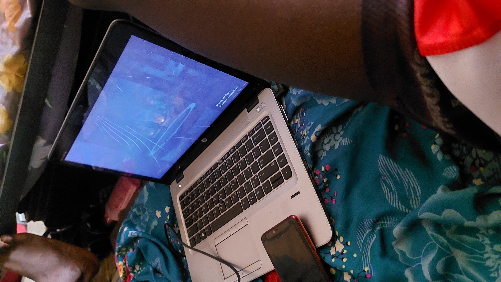 A low-angle, close-up shot captures a laptop on a bed with teal, floral patterned sheets. The laptop screen is cracked and displays a blue background with a "Activate Windows" prompt. A red-cased smartphone rests to the left of the laptop, partially covering the touchpad. The scene appears to be indoors, possibly in a bedroom, in New Nyanya, Nigeria. The presence of a foot in the upper left corner suggests a person is present but not fully visible. The overall lighting is dim, contributing to a relaxed or casual atmosphere.