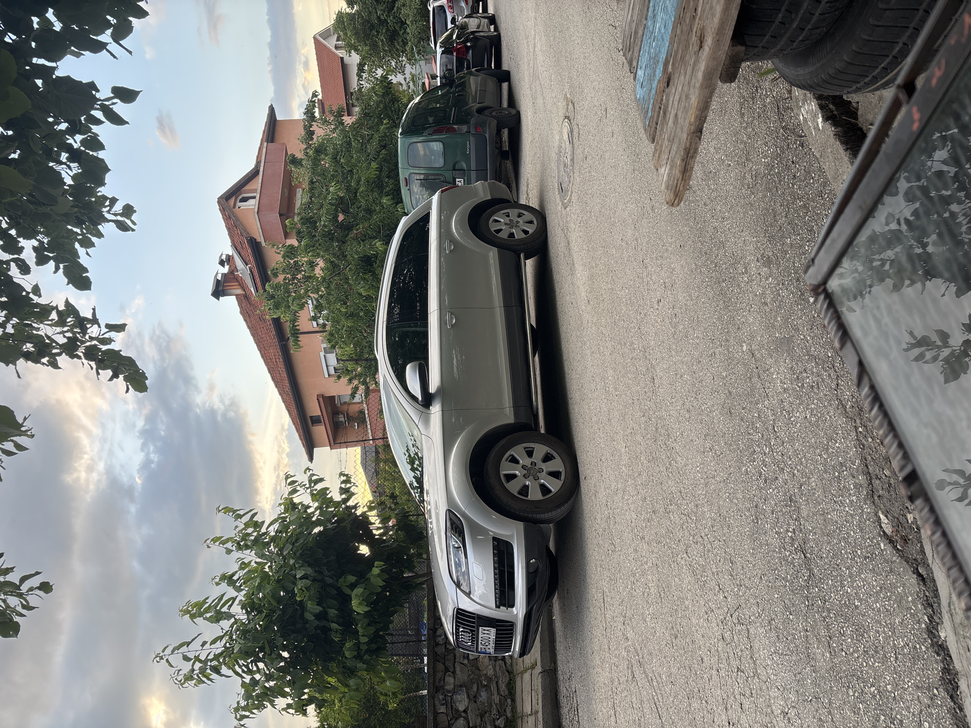 The image shows a silver Audi Q7 parked on a narrow street in Asenovgrad, Bulgaria. The street appears to be residential, with a multi-story building visible in the background. Several other cars are parked further down the street. On the right side of the frame, there are tires, wood and part of a mirror. The sky is cloudy. The angle of the photograph is slightly tilted.