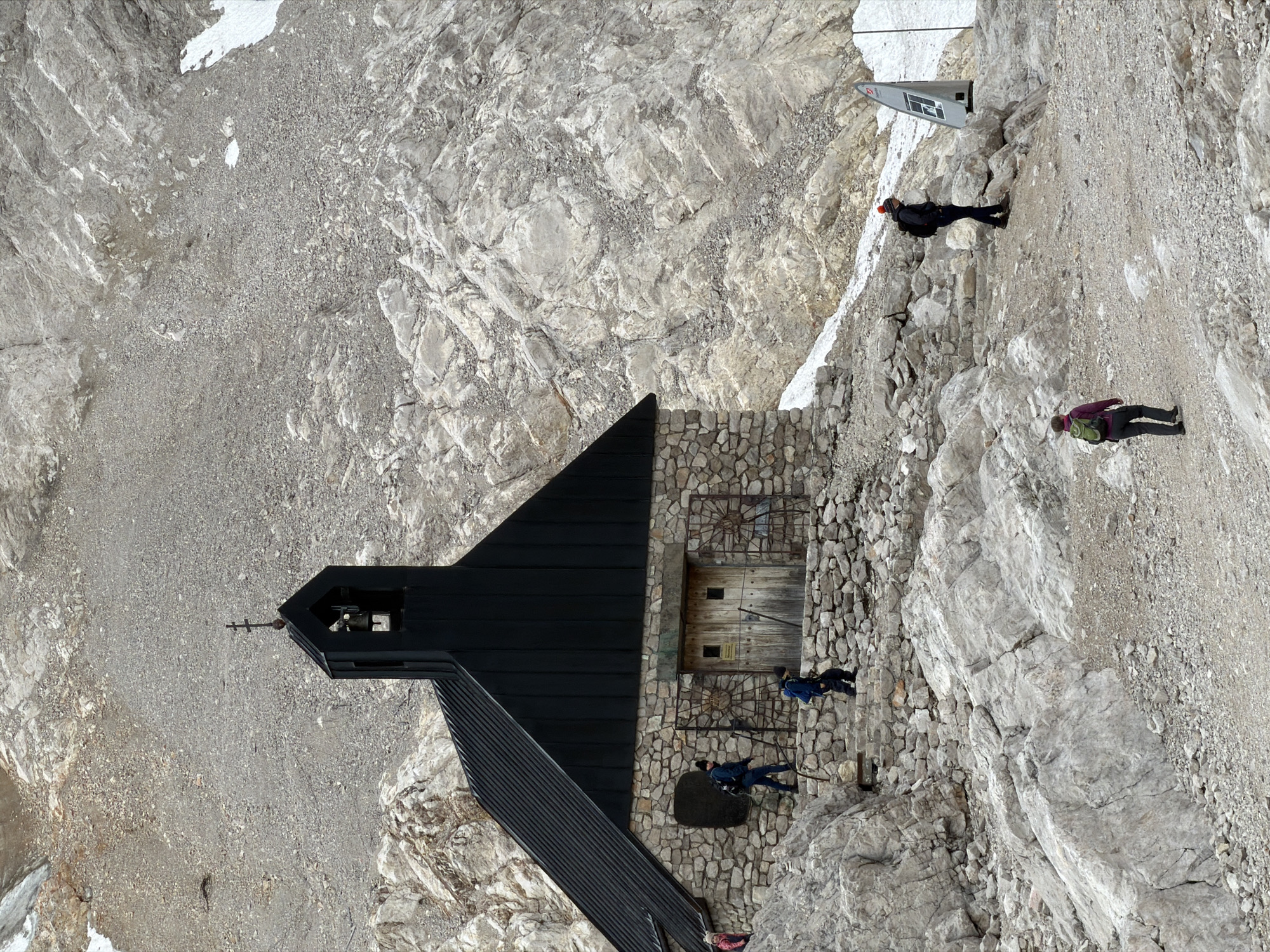 The image depicts the small chapel known as the "Mariä Himmelfahrt" (Assumption of Mary) chapel, located near the summit of the Zugspitze mountain, Germany's highest peak, situated in the municipality of Garmisch-Partenkirchen. The chapel is constructed of stone with a dark, pitched roof and a small bell tower topped with a cross. Several individuals are present in the scene, walking on rocky terrain and a stone staircase leading up to the chapel. One person in the foreground is walking away from the camera with a green backpack, while another person in the mid-ground wears a red beanie and a dark jacket, looking at a signpost. Two individuals are ascending the stone steps towards the chapel, one in a blue jacket and the other in a dark outfit. The environment is mountainous, characterized by large rock formations, gravelly ground, and patches of snow, indicative of a high-altitude alpine setting.
