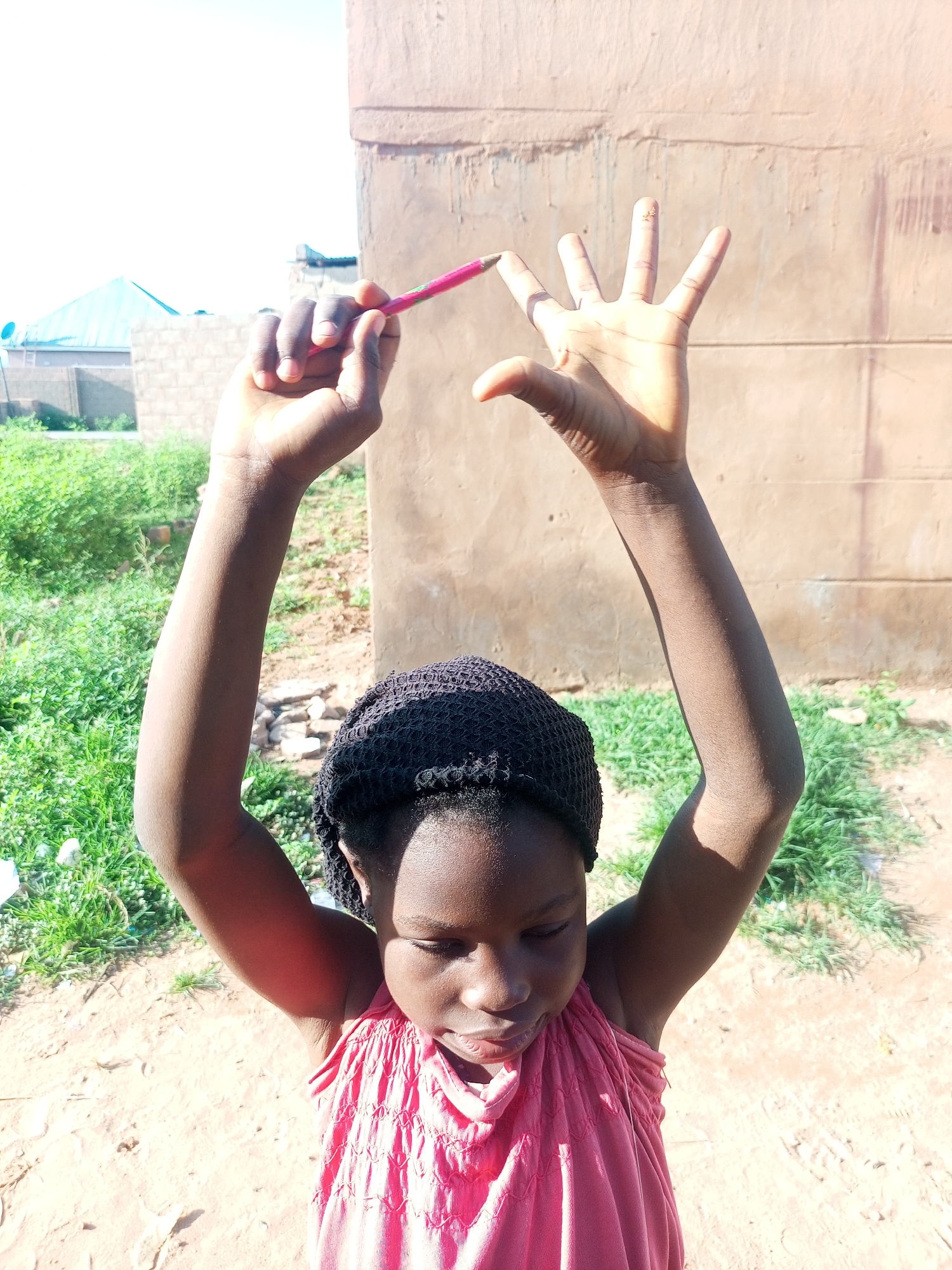 A young girl, wearing a pink top and a black head covering, stands outdoors in what appears to be a residential area in Tempure, Nigeria. She is holding a pink pencil in her right hand, positioned in front of her face, and her left hand is outstretched with fingers spread. The background consists of a rough, textured wall, some green vegetation, and a dirt or sandy ground. The sky is visible and appears bright, suggesting daytime. The overall scene captures a moment of playful engagement with the pencil and a gesture of open hand.