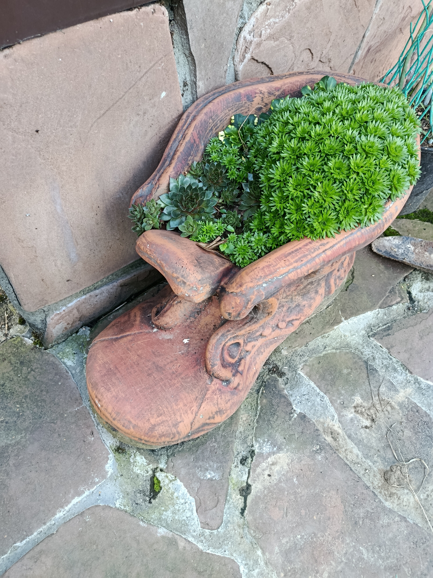 A terracotta planter, shaped like a human face, is filled with succulents and moss. The planter is positioned on a stone patio with cobblestone and flagstone elements. A portion of a building's foundation, made of a rough, brown stone, is visible behind and to the left of the planter. A green metal fence is present in the upper right background. The image appears to be an outdoor scene in a garden or yard setting. The location is specified as Misaylovo, Russia.