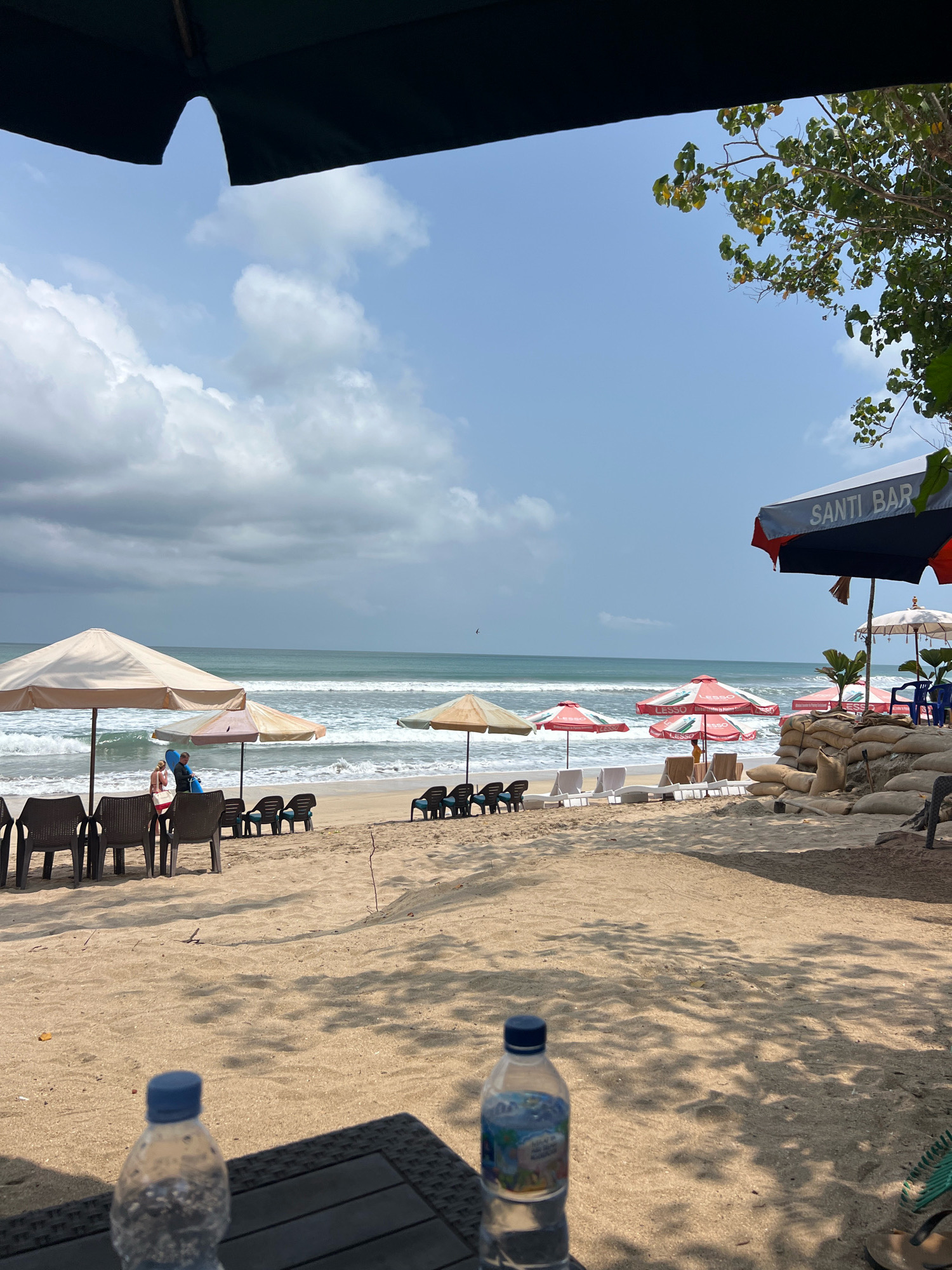 The image depicts a beach scene in Kuta, Indonesia, on a cloudy day. In the foreground, a table holds two plastic water bottles, one in focus and the other slightly blurred. The table is casting shadows on the sand. Further back, beach chairs and umbrellas are set up on the sandy shore. Some of these chairs are unoccupied, while a couple of people are standing near umbrellas. The ocean stretches out behind the chairs, with moderate waves. A few sandbags are piled near some of the umbrellas, possibly for flood defense. A sign on one of the umbrellas reads "SANTI BAR". Trees are visible on the right side of the image, partially obscuring the sky. The overall atmosphere suggests a relaxed beach setting, potentially a commercial establishment on the beach.